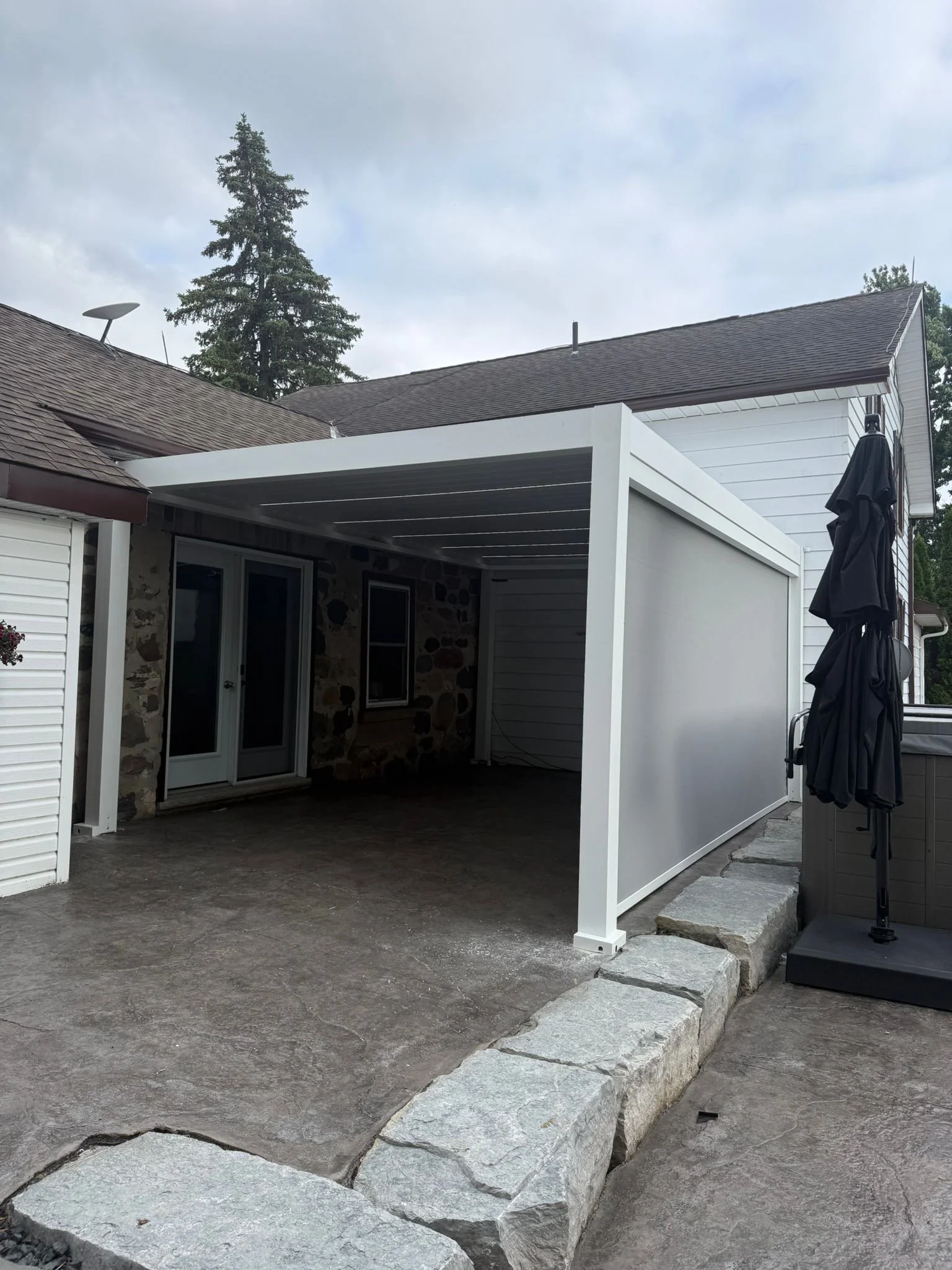 Backyard patio with concrete floor, stone border, attached white pergola, sliding glass doors, and a black patio umbrella.