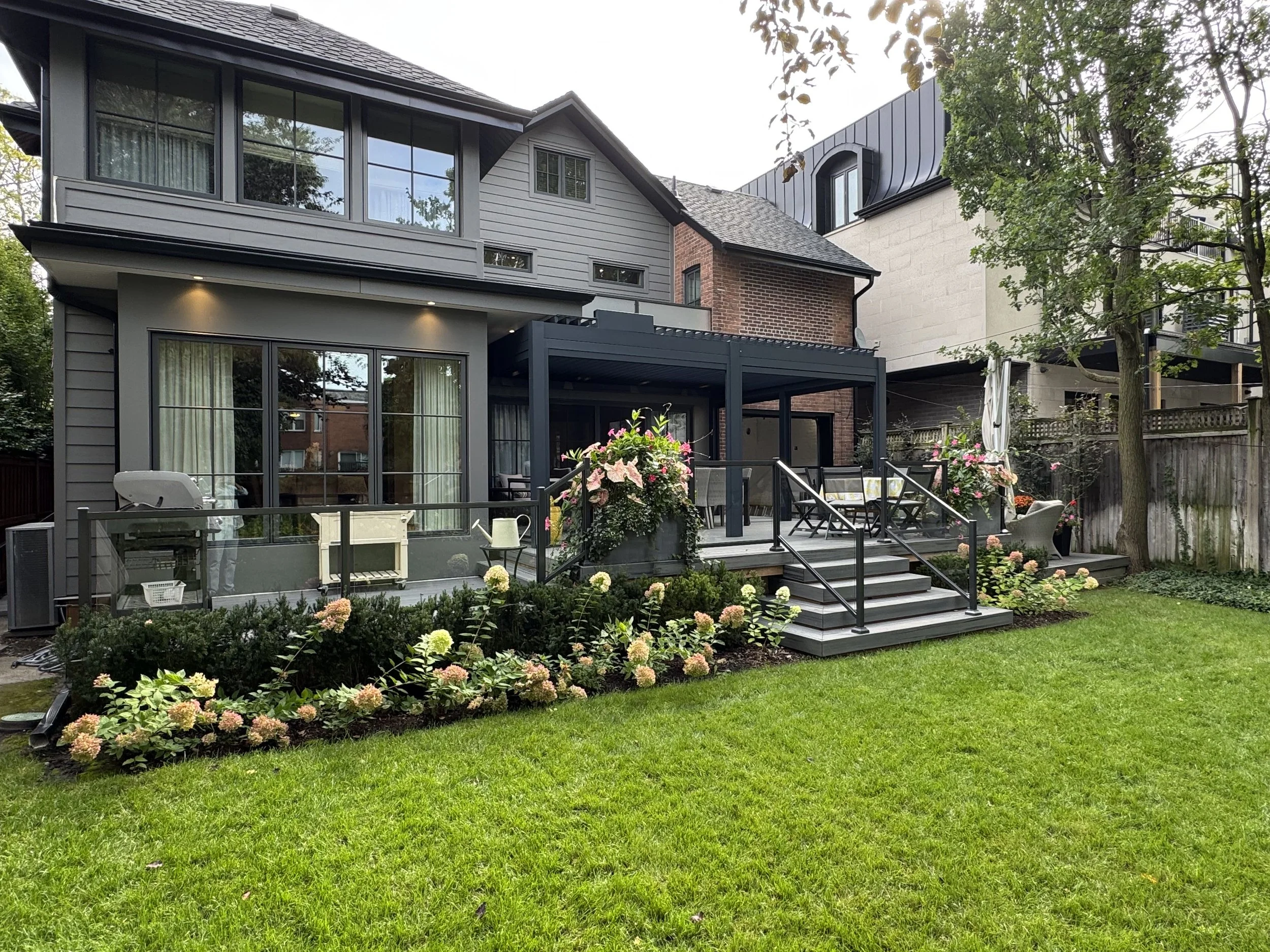 A backyard outdoor patio area with a modern two-story house in the background. The patio has a table with chairs, decorative flowers, and some gardening tools and supplies. The house features large windows, a mix of gray and brick exterior, and a bla