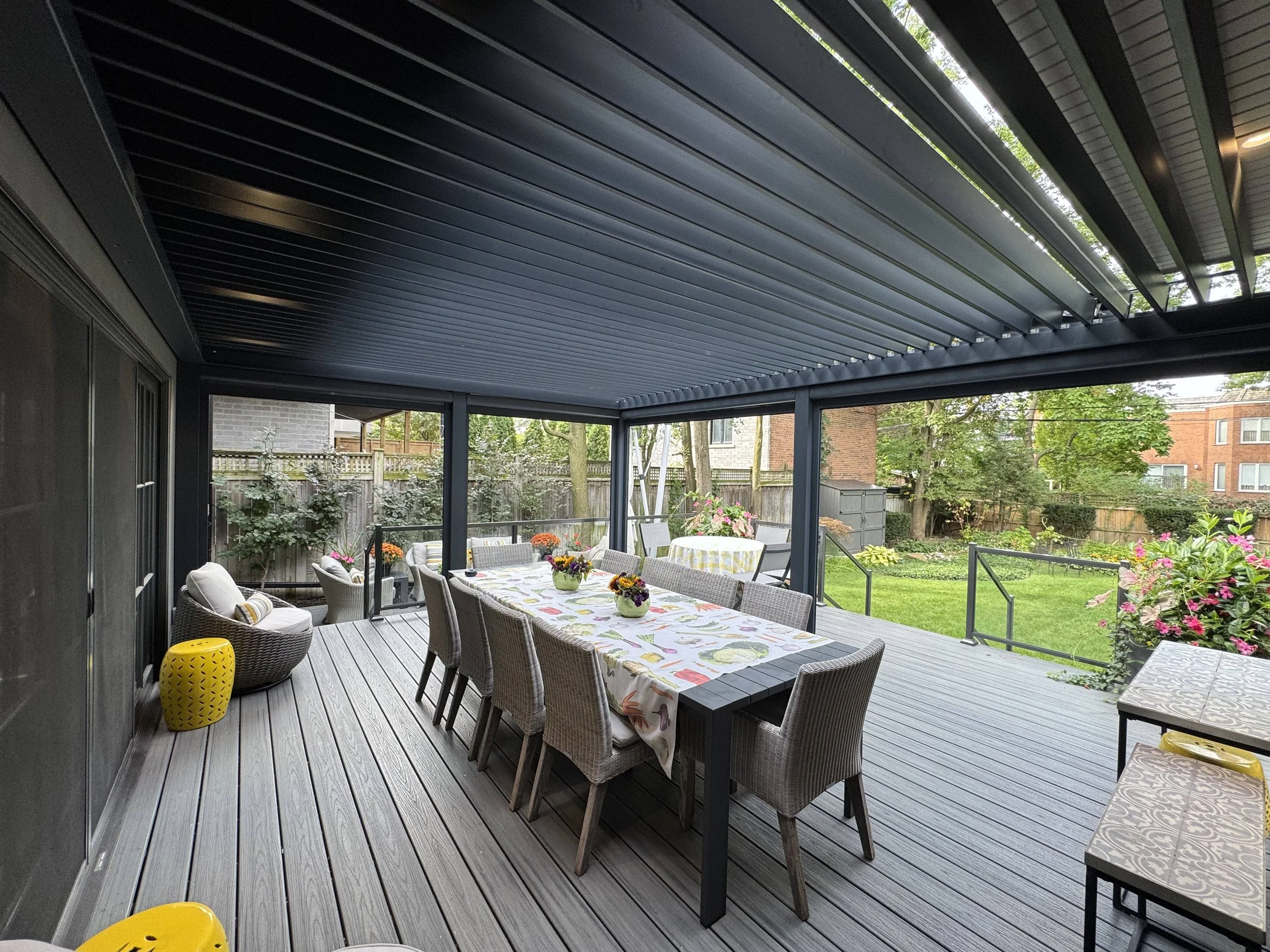 Covered patio with a dining table covered with a tablecloth, surrounded by wicker chairs with cushions, overlooking a backyard garden with trees and flowers.