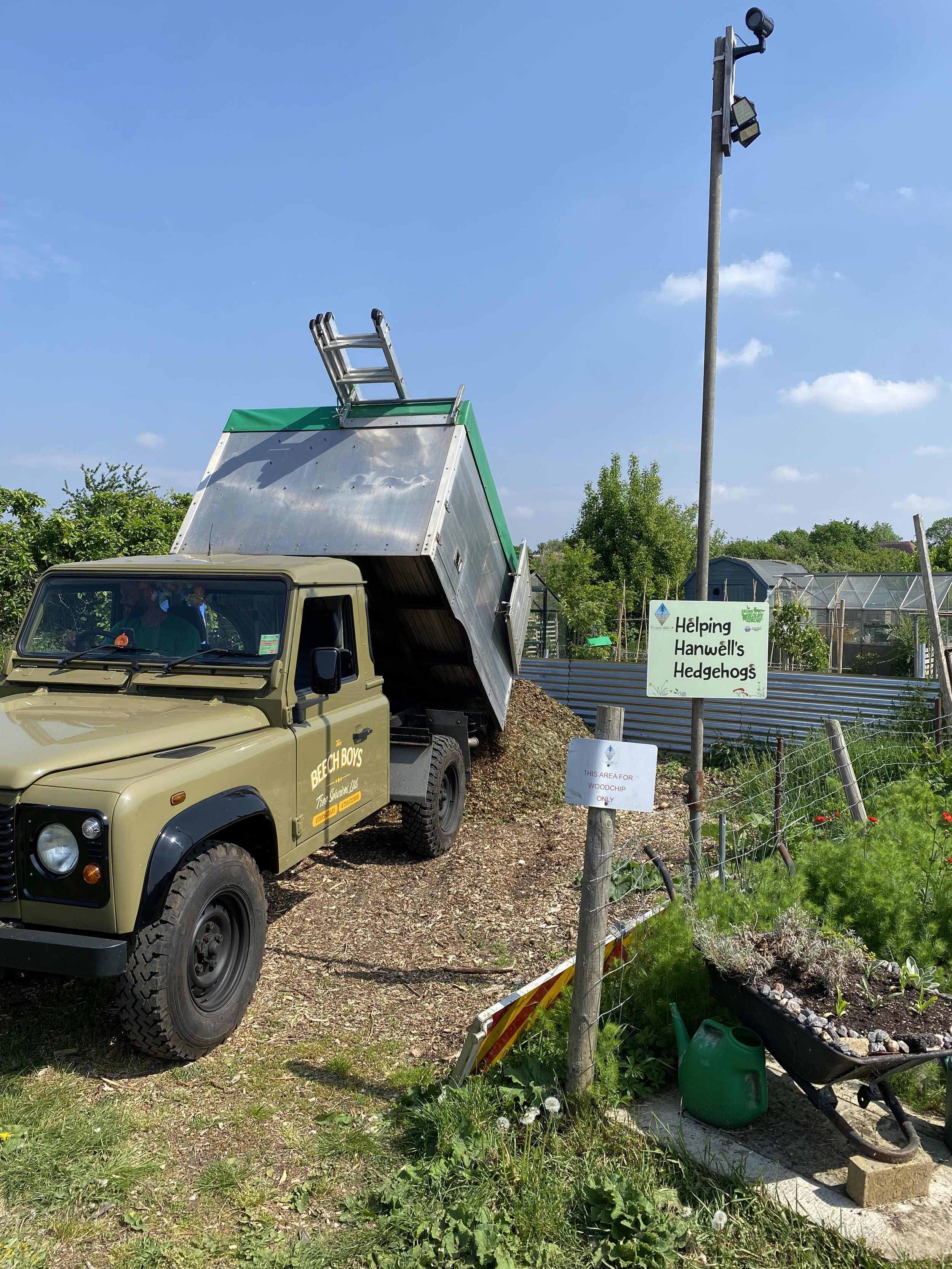 A small truck dumping soil or compost at a community garden with signs indicating it is for helping Hanwell's hedgehogs and a specific area for woodchipping. The truck is in a rural setting with green plants, a wheelbarrow, and gardening supplies nearby.