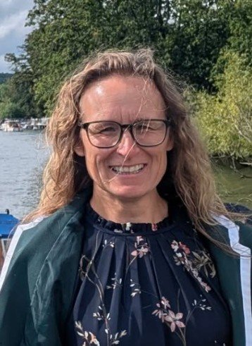 English tutor in dark blue floral dress and a green blazer standing by a river and trees