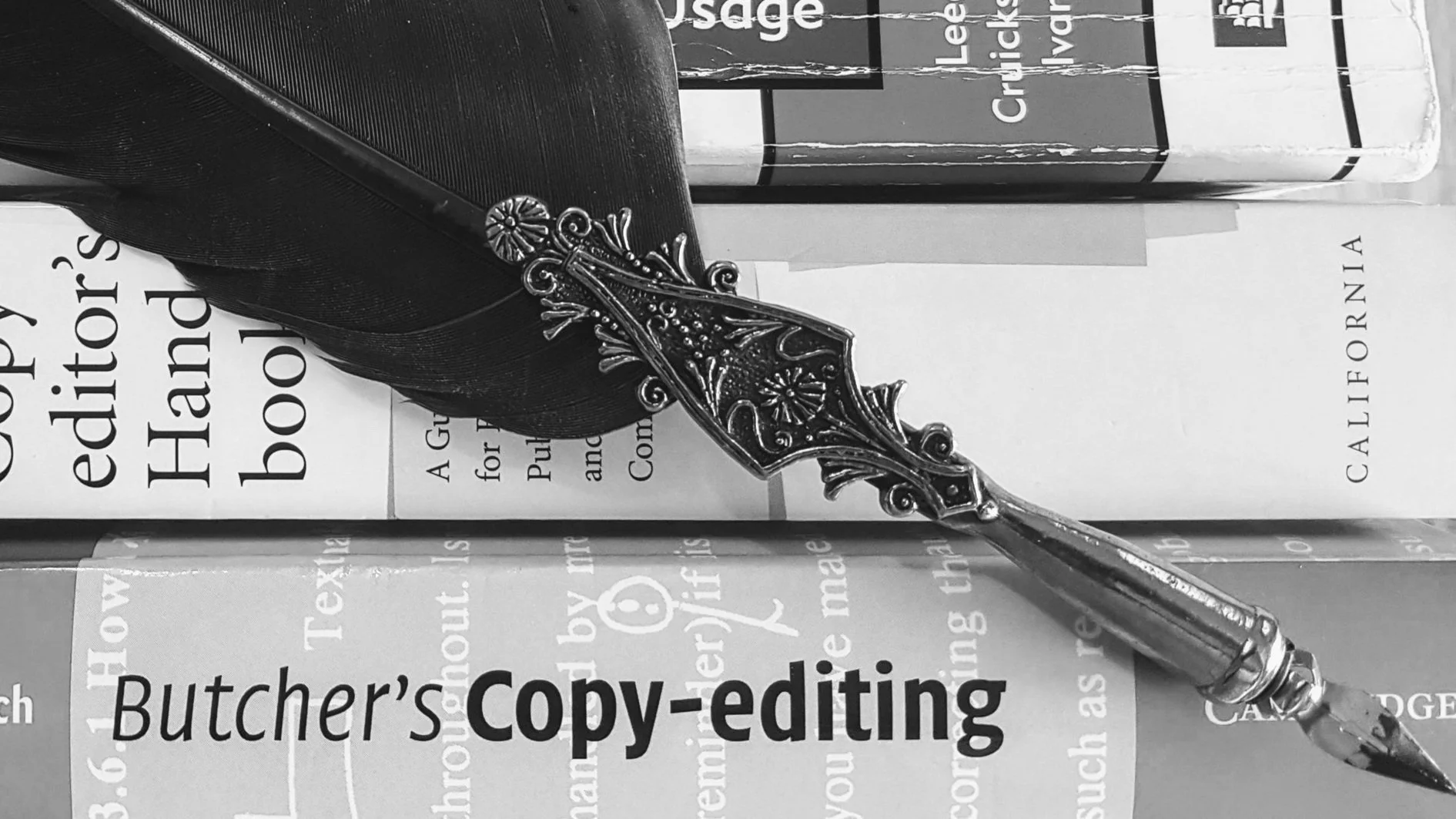 A black feather quill pen with an ornate metallic decorative end, resting on several books and papers including one titled 'Butcher's Copy-editing' and another that mentions 'Elements of Style'.