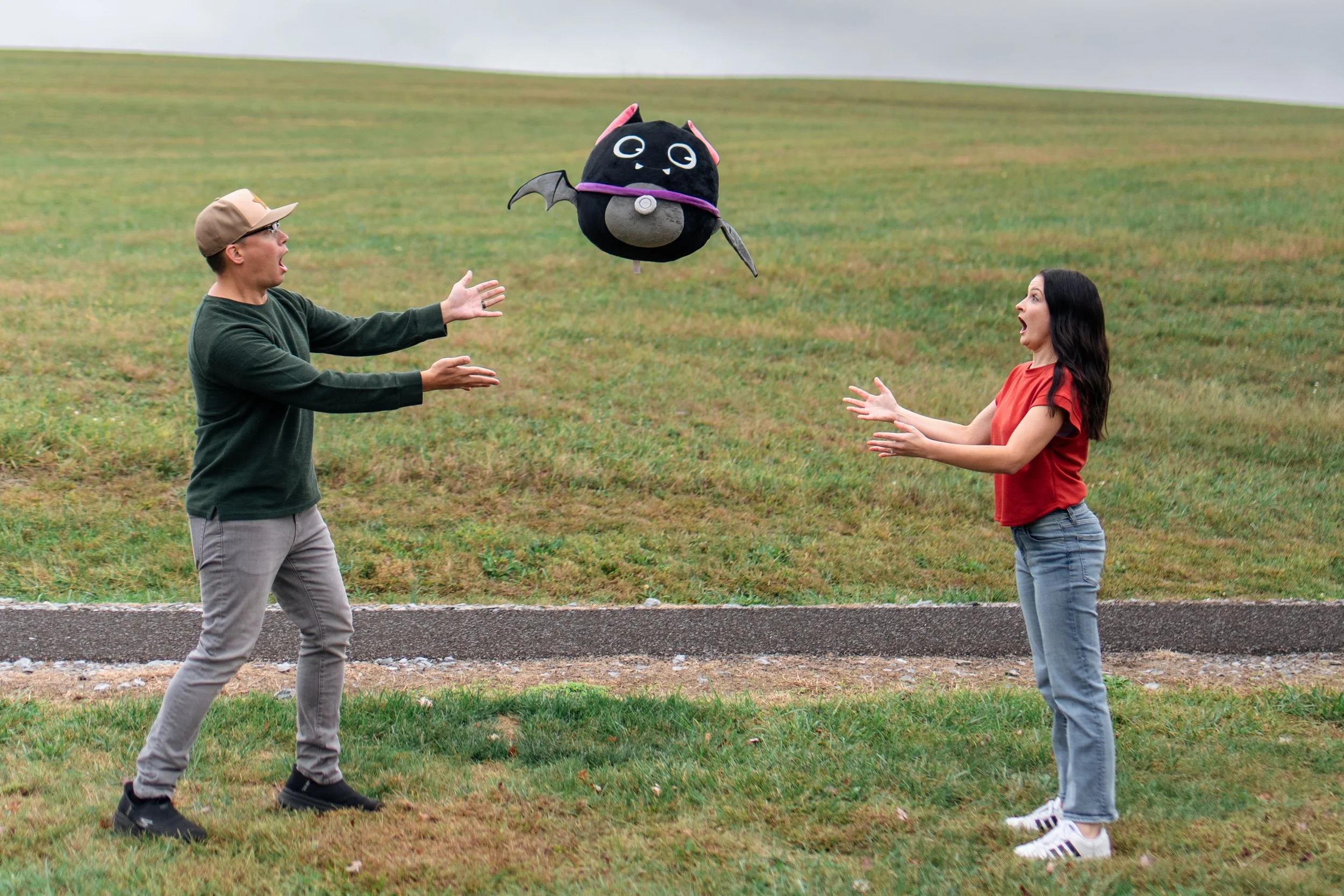 A man and woman standing on a grassy area, both with surprised expressions, as a large black bat-themed drone with eyes, wings, and a bat-shaped design flies between them on a cloudy day.