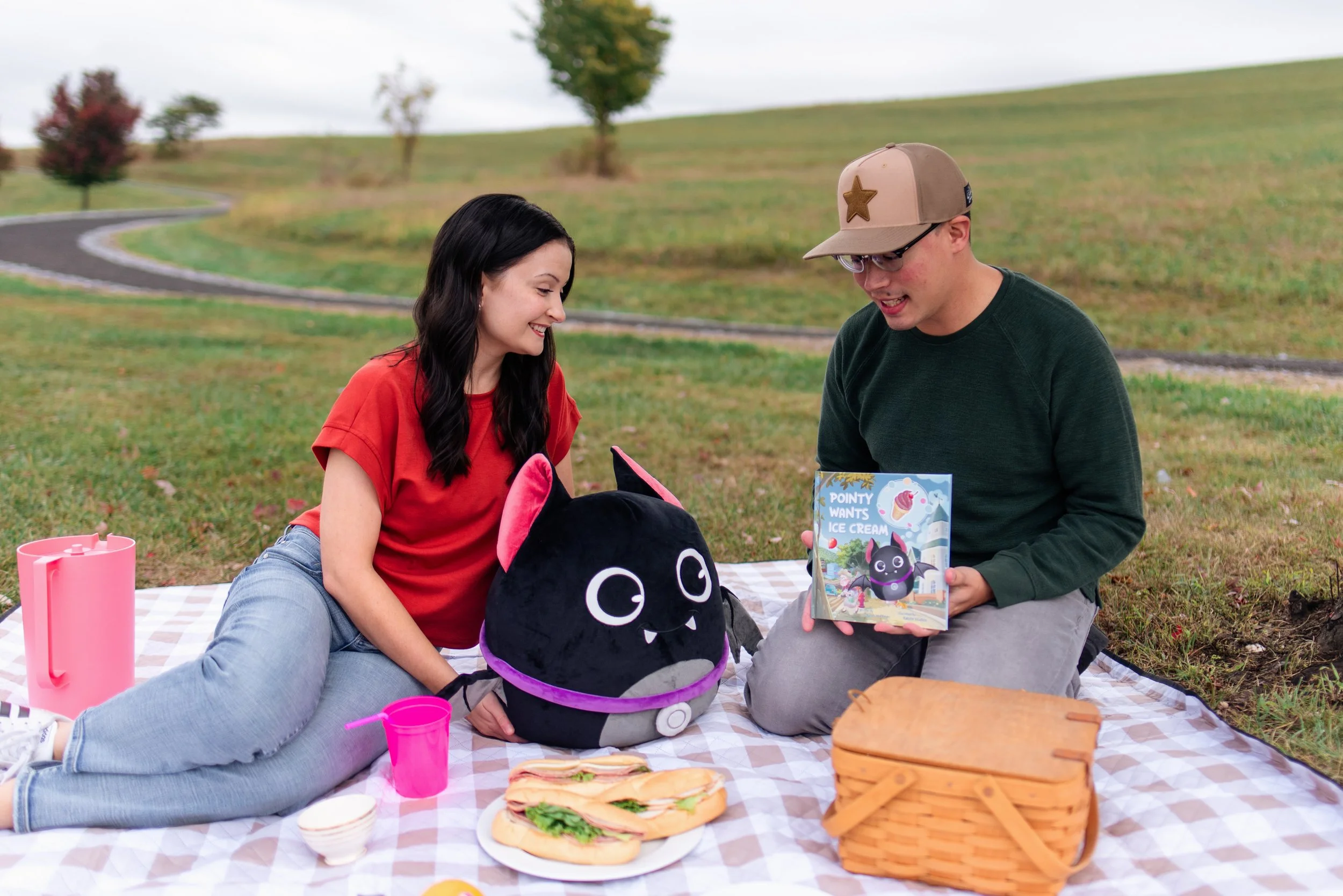 A young woman and man having a picnic outdoors on a checkered blanket. The woman has black hair, and the man has glasses, a cap with a star, and a green shirt. They are smiling at each other, with snacks like sandwiches and a pink thermos around them. A plush backpack and a picnic basket are also visible.