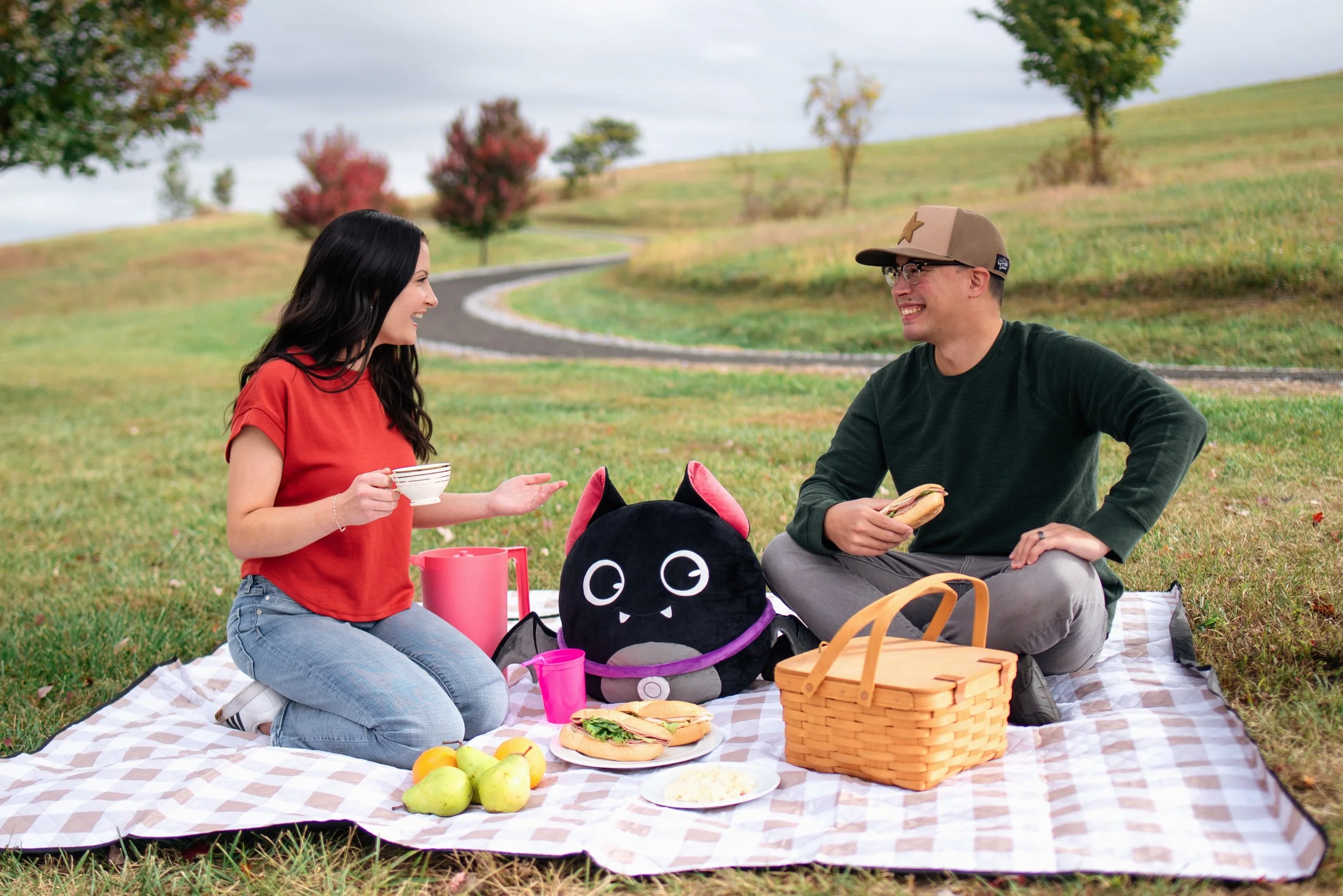Two people having a picnic on a checkered blanket in a park with trees and a winding path, with a plush black cat bag and picnic food including sandwiches, apples, and drinks.