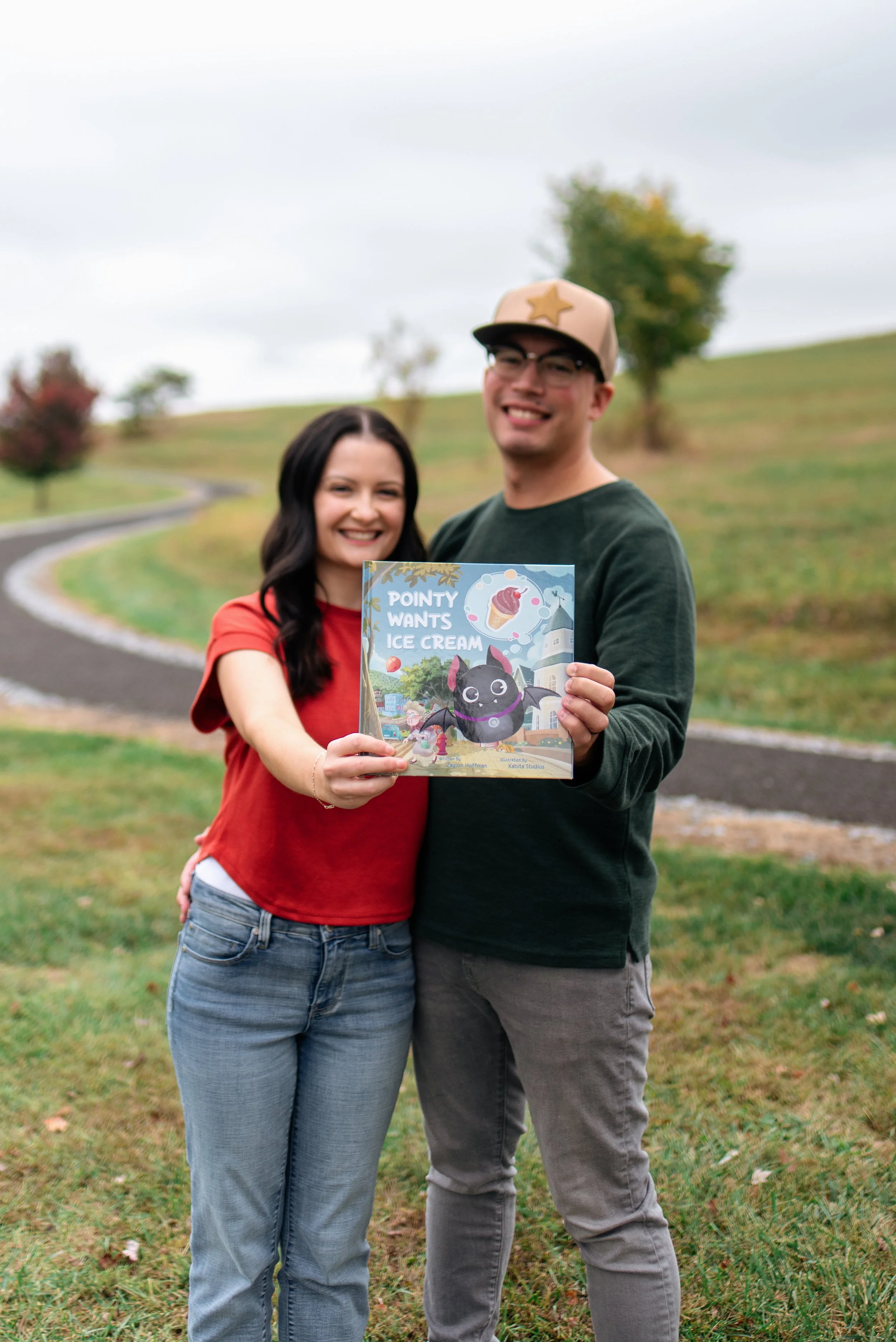 A smiling woman with dark hair and a man with glasses and a beige cap holding a children's book titled "Pointy Wants Ice Cream" outdoors on a grassy hill with trees and a winding path in the background.