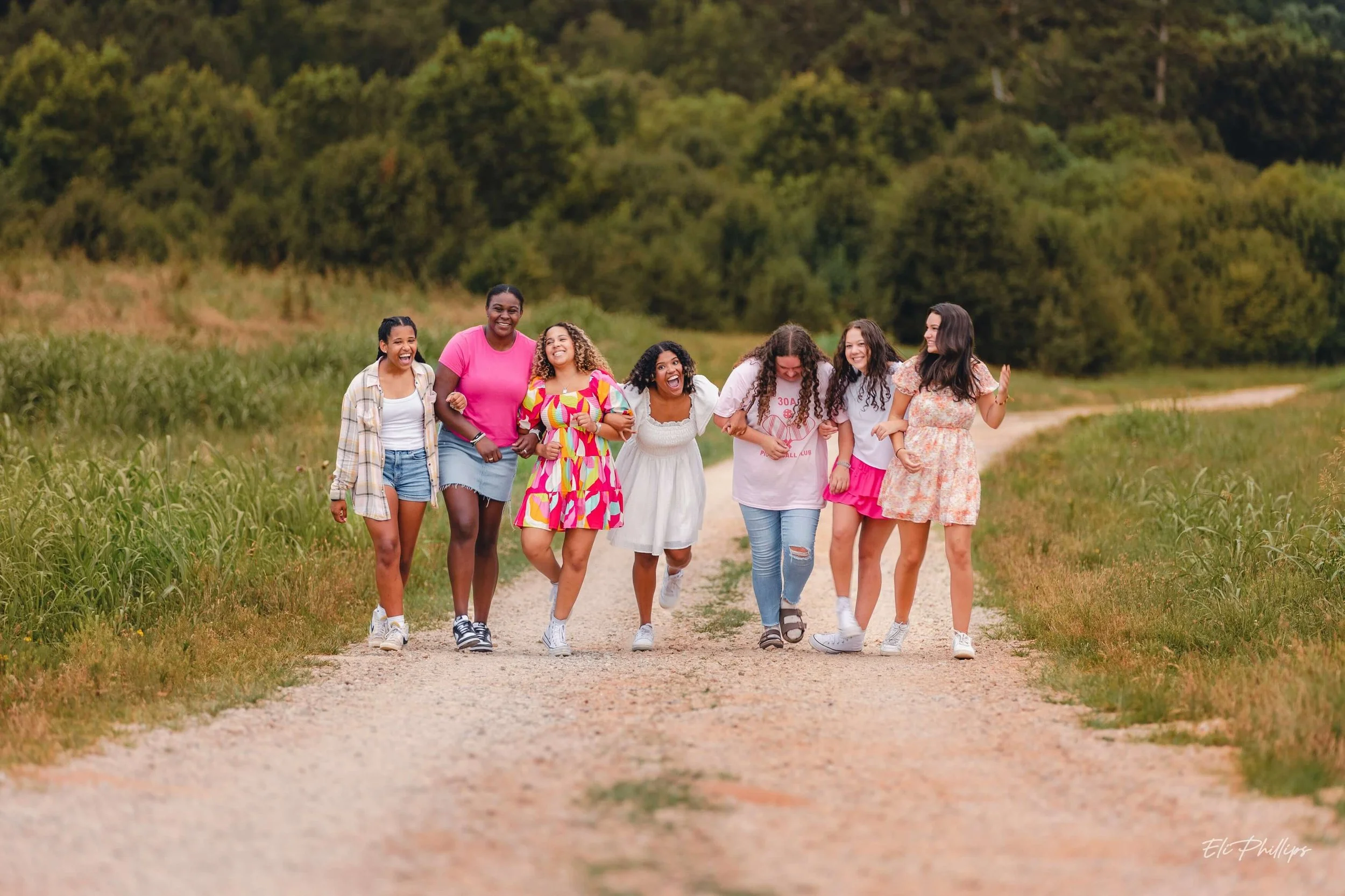 Group of seven women walking arm-in-arm on a dirt path through a grassy field with trees in the background, smiling and laughing.
