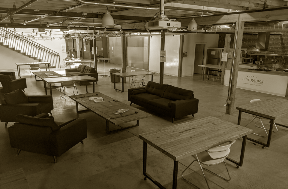 An industrial-style office space with black sofas and chairs, wooden tables, a staircase on the left, and a kitchenette in the back right. The space has exposed pipes and brick walls.