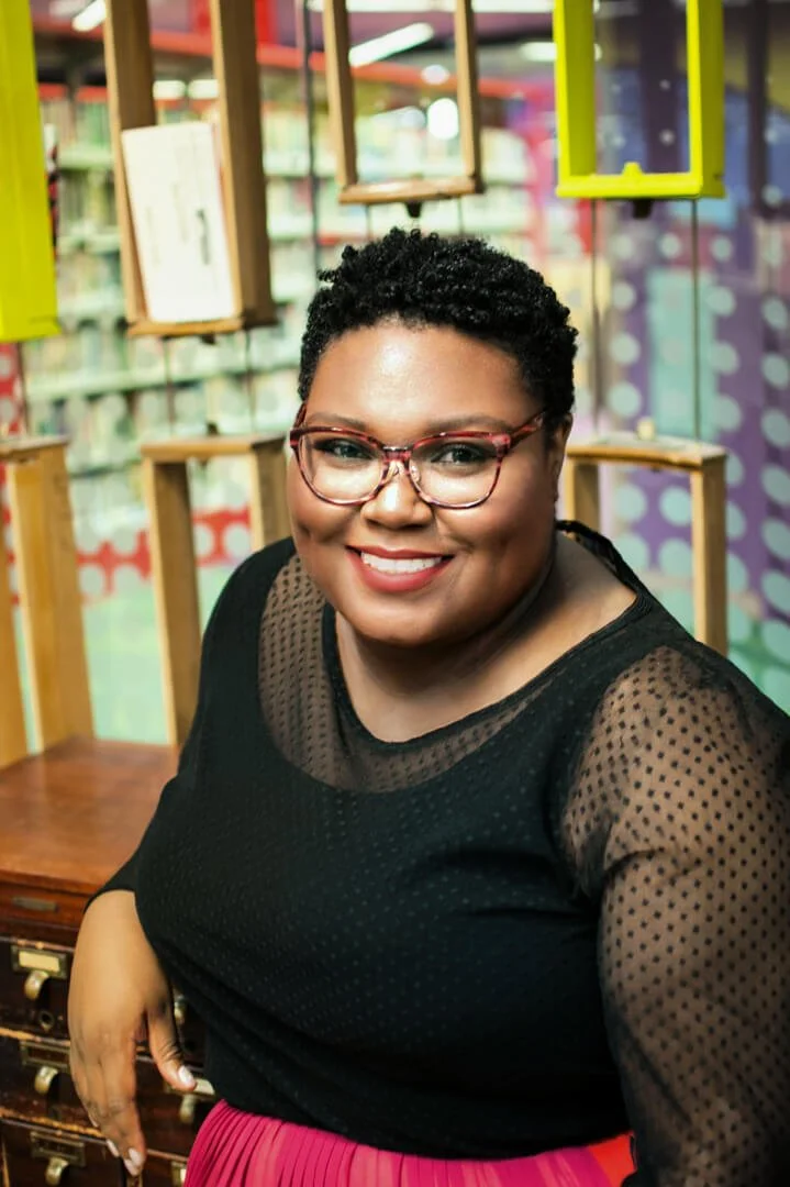 Rhodesia Jackson with glasses smiling in a colorful indoor setting.