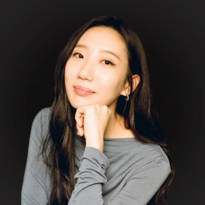 Irene Song with long dark hair wearing a gray top, resting her chin on her hand, smiling gently, against a black background.
