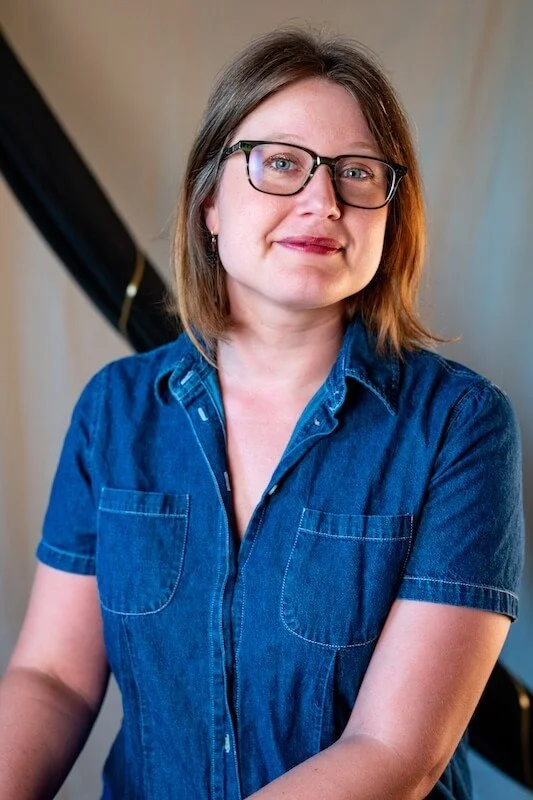 Laura Nash with shoulder-length brown hair and glasses, wearing a blue denim shirt, standing indoors.
