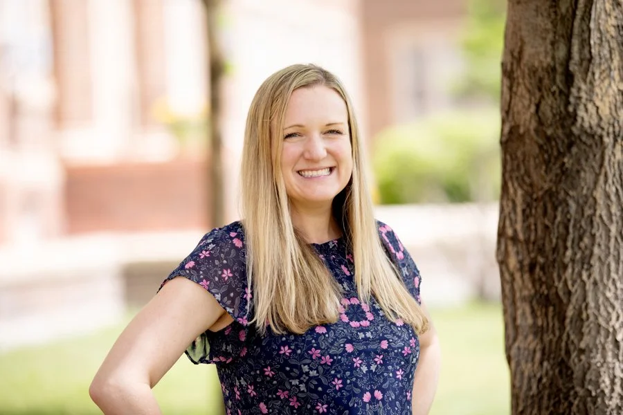 Brittany Cowan with long blonde hair wearing a navy blue floral dress, smiling outdoors next to a tree with a blurred background of greenery and buildings.