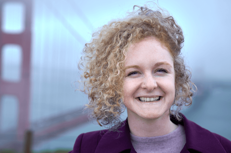 Ashley Kochans with curly blonde hair smiling outdoors near the Golden Gate Bridge in San Francisco.