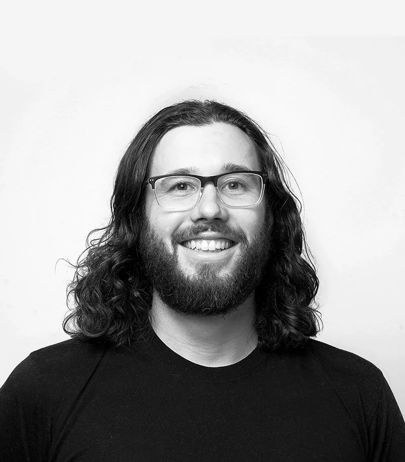 Beren Davis with long curly hair, glasses, and a beard smiling in a black shirt against a plain background.