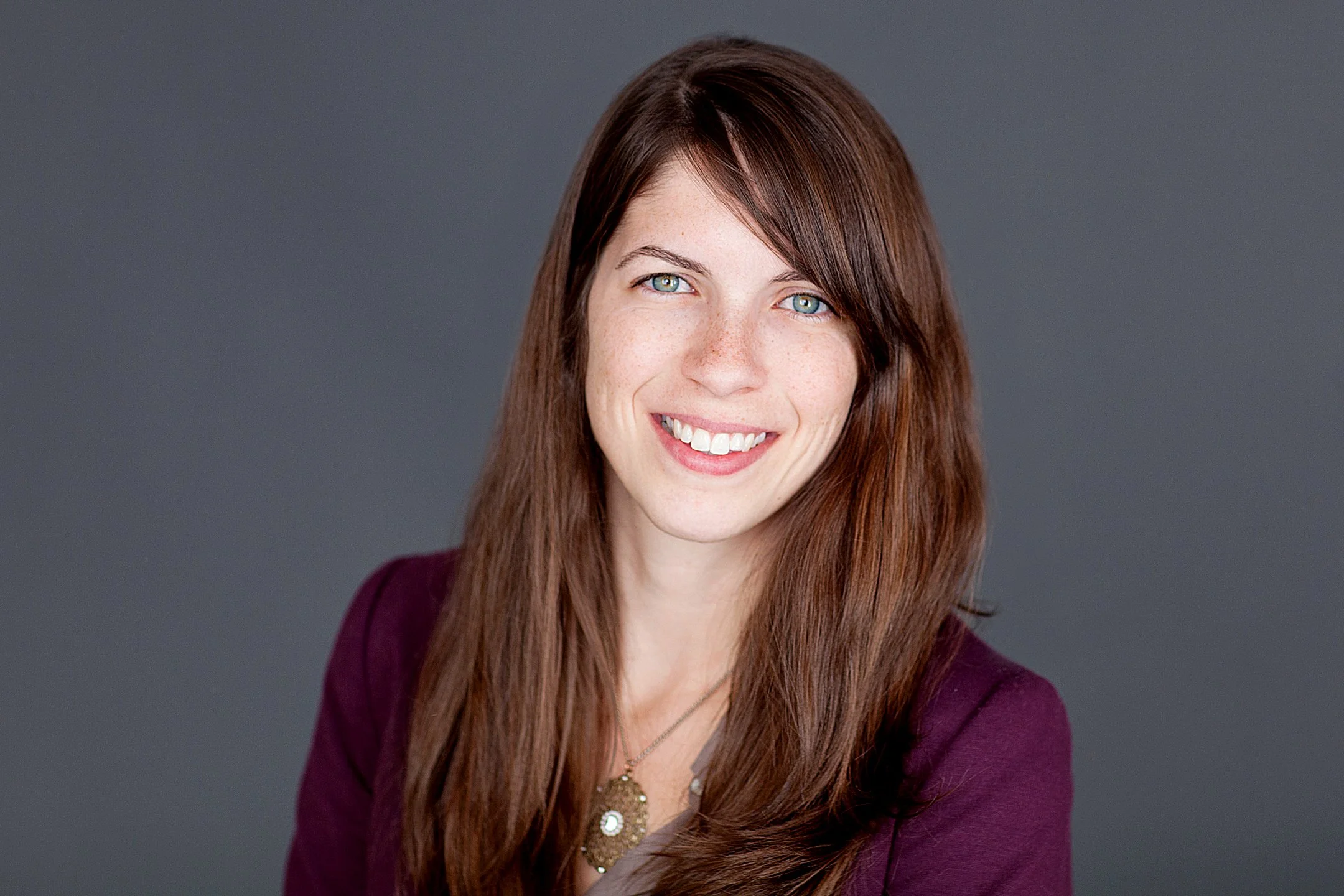 Annetta Crecelius smiling with long brown hair, blue eyes, wearing a maroon blazer and a necklace, against a dark gray background.