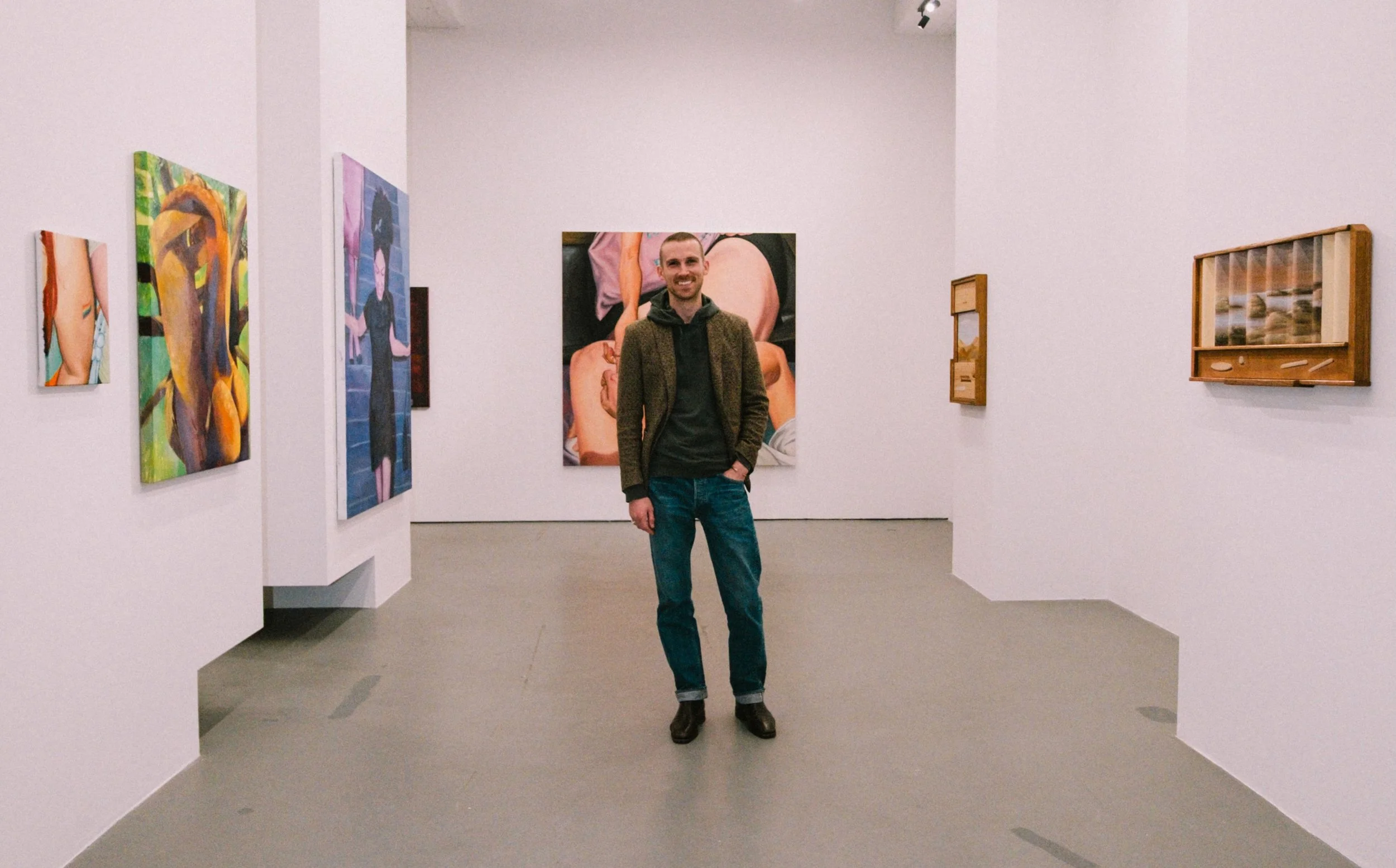A man in a brown jacket, black hoodie, and jeans stands smiling in an art gallery with various colorful paintings on white walls.