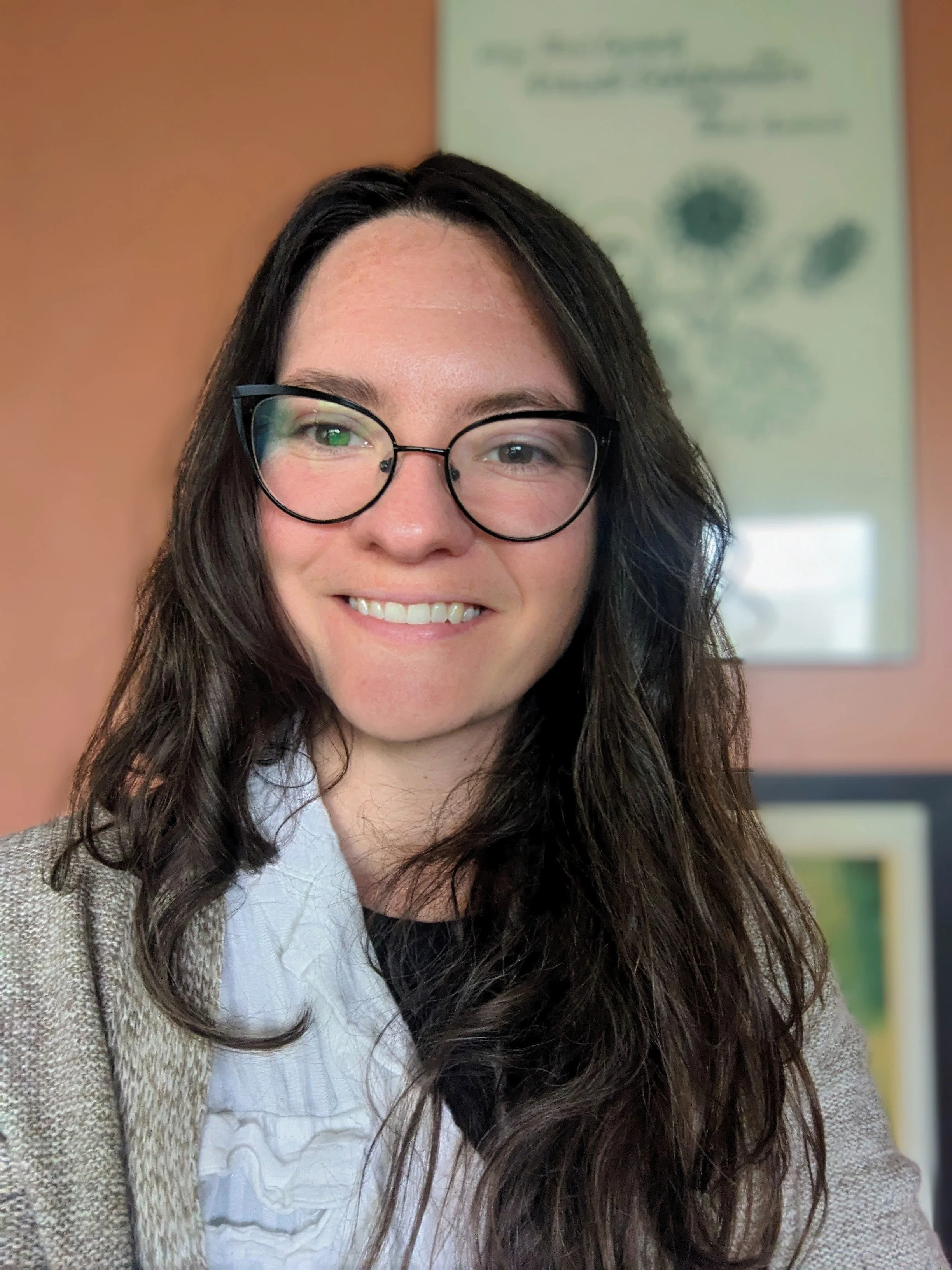Elise, A woman with long dark hair, glasses, and a warm smile, in her office with with framed pictures and a plant poster on the wall behind her.