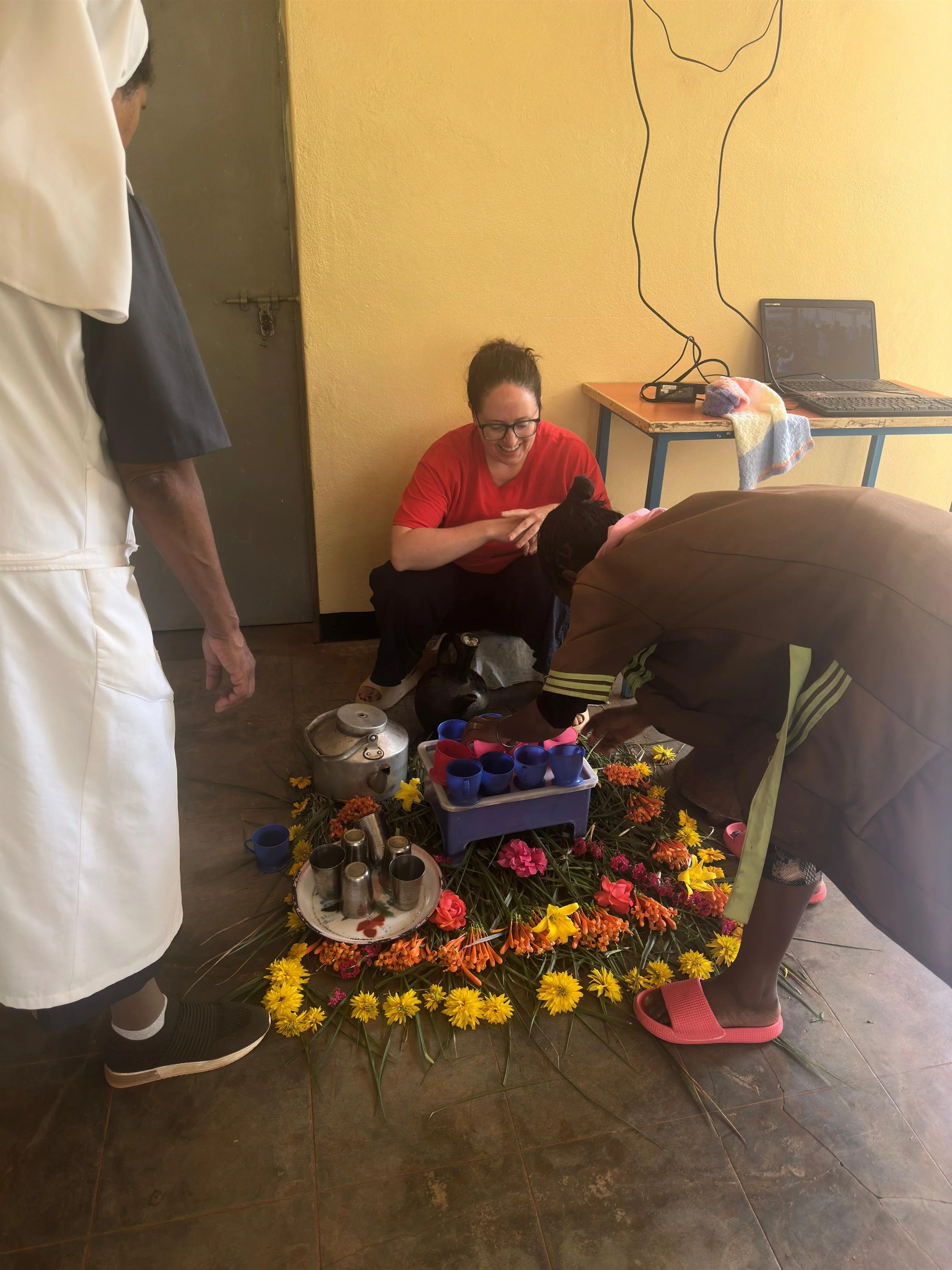 A woman in red sitting on the floor, smiling, participating in a traditional ceremony with a woman pouring liquid into small cups arranged on a tray on the floor surrounded by colorful flowers. A person in a brown jacket and pink slippers is leaning over the setup, and there is a person in white and another in white and black clothing standing nearby. A table with a laptop, a towel, and some cables is in the background.