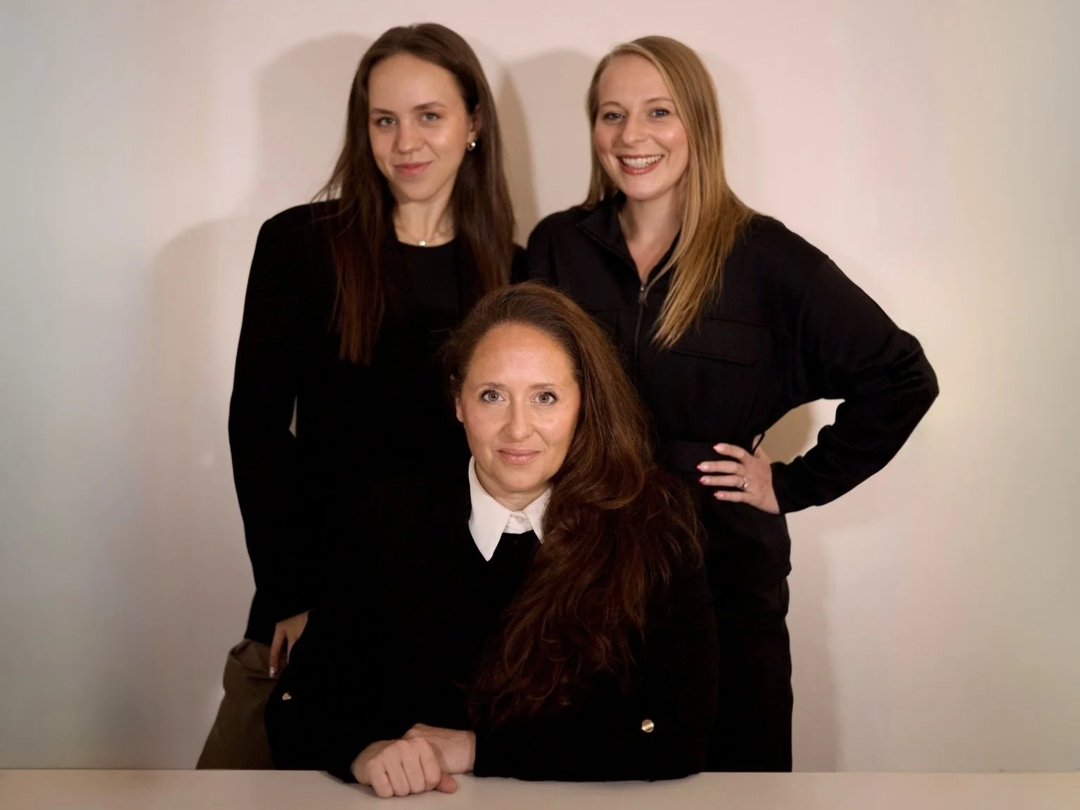 Three women posing together for a photo in front of a plain background, two standing behind and one seated in front.