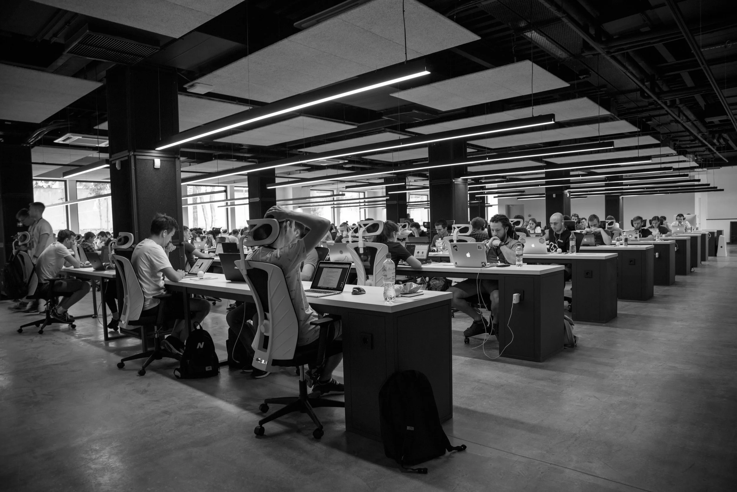 A large modern coworking space or open office filled with many people working at desks with laptops and water bottles, illuminated by overhead lighting.