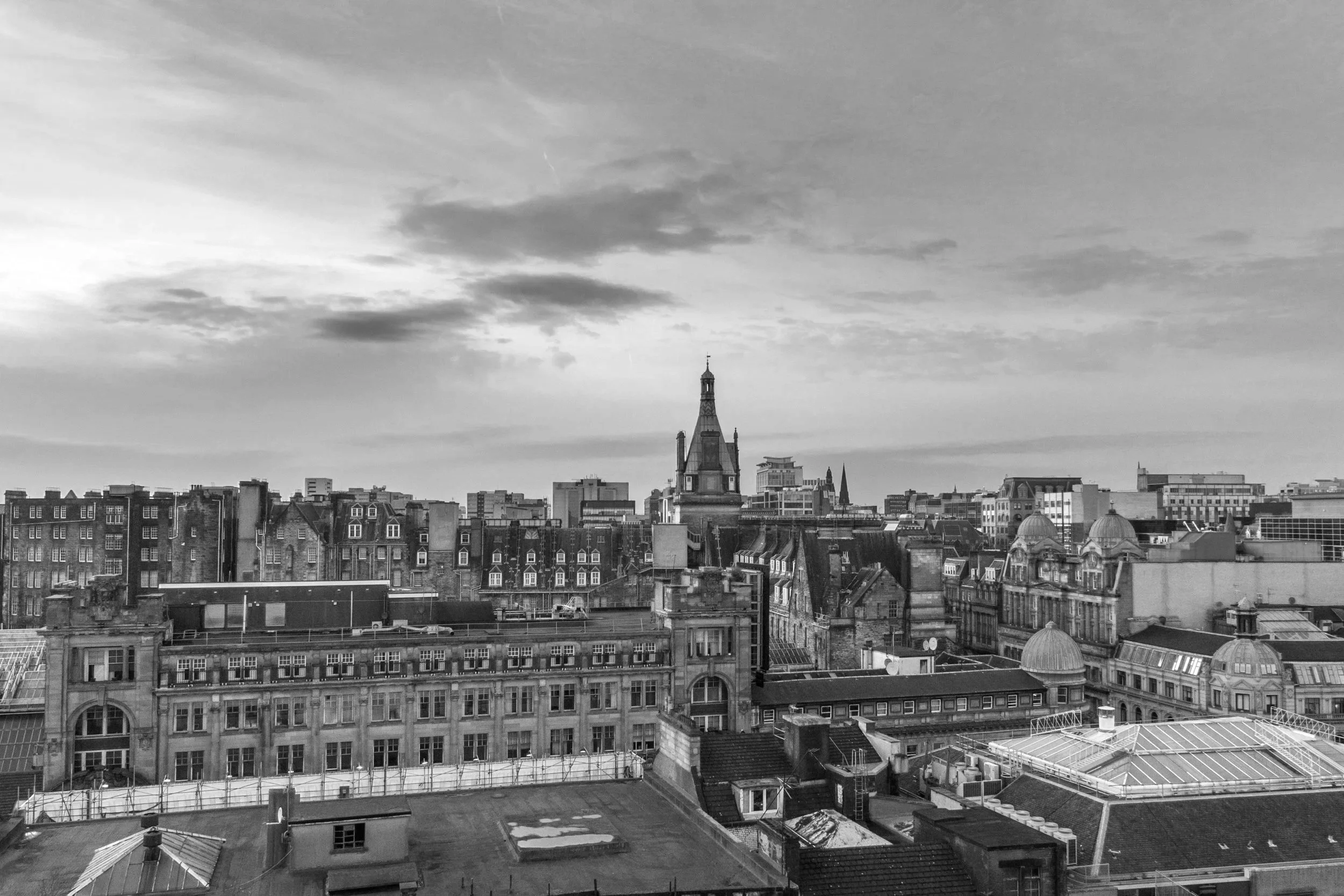 Black and white aerial view of a city skyline with a mix of historic and modern buildings, and a prominent church tower in the center under a cloudy sky.