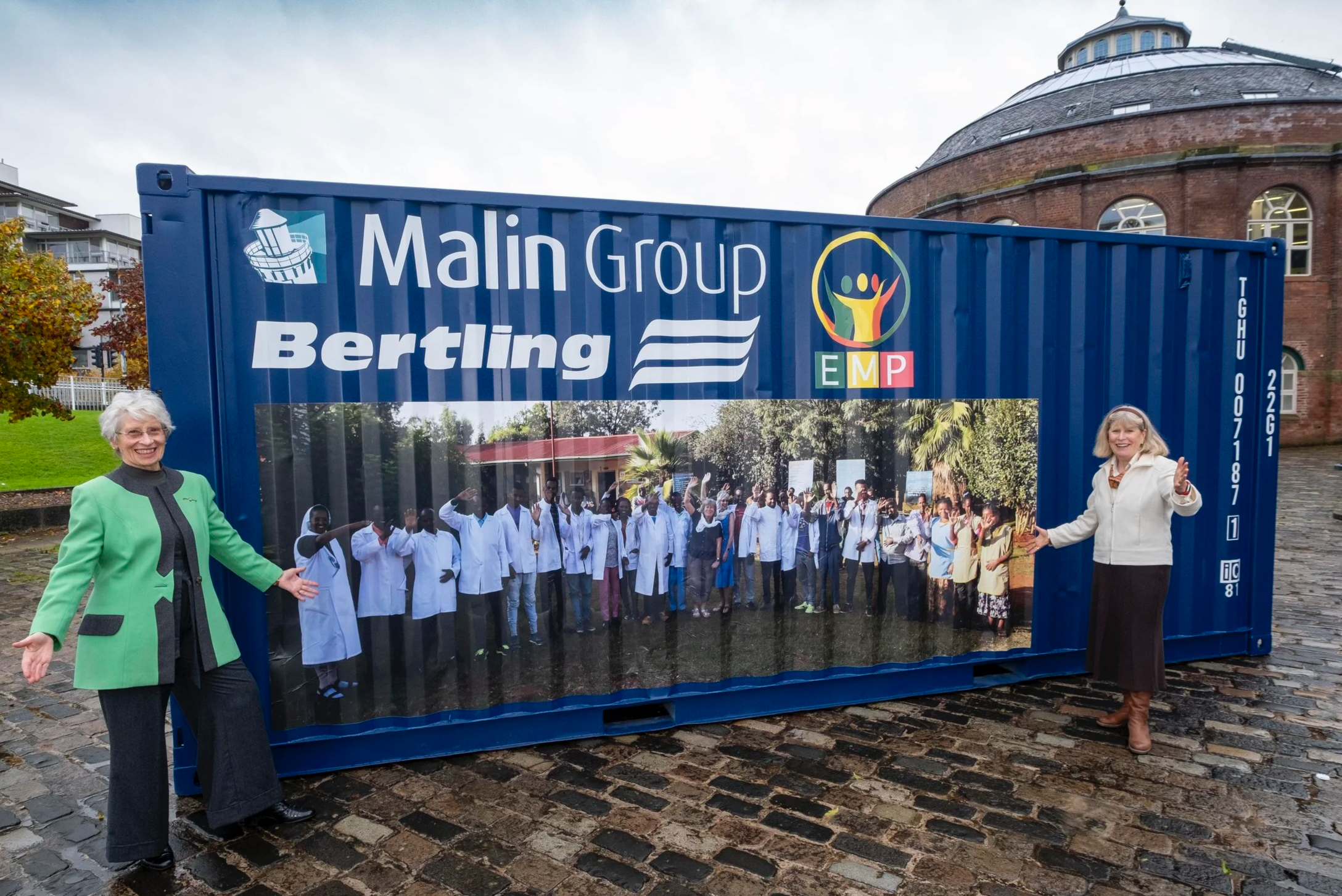 Two women standing on either side of a large container with a group photo and logos on it. They are outdoors on a cobblestone street, smiling and waving.