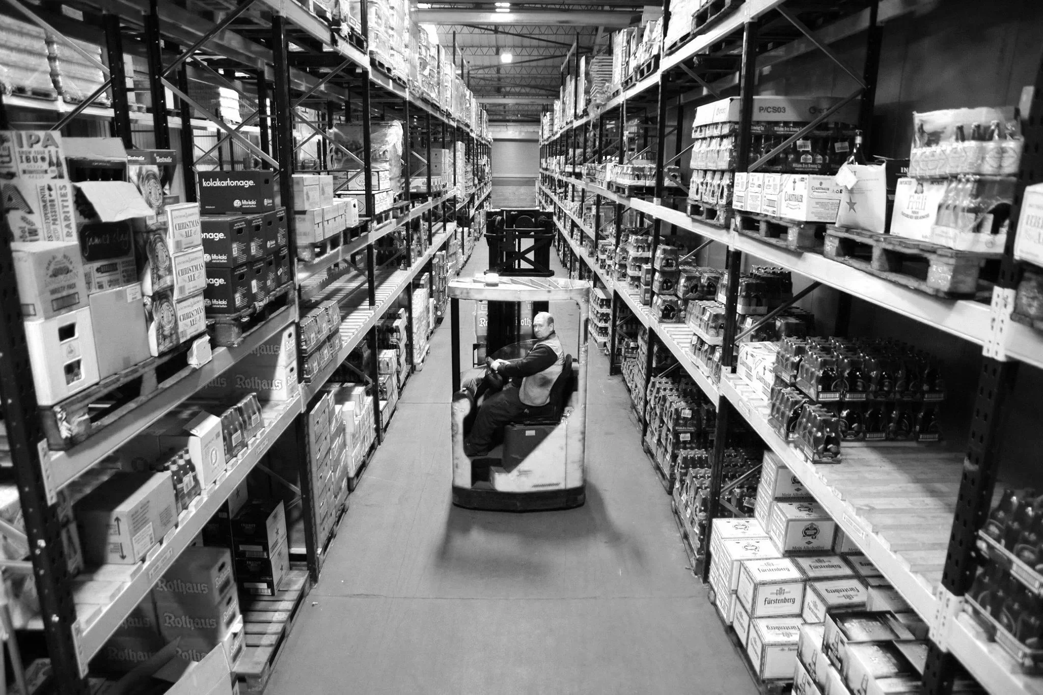 A warehouse aisle with tall metal shelving filled with boxes and products on both sides. A man is operating a ride-on pallet truck in the center of the aisle, looking back towards the camera.