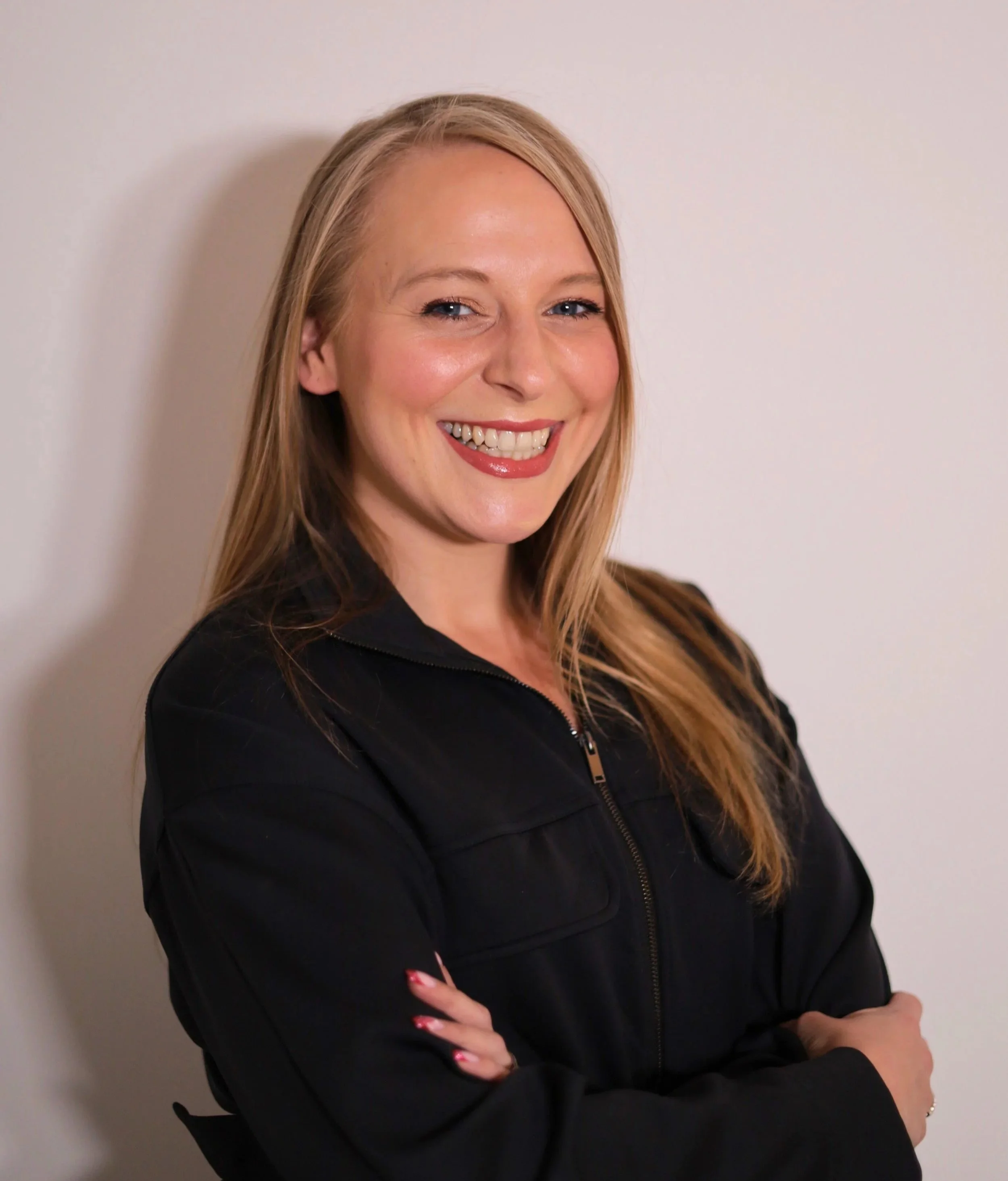 A woman with long blonde hair, smiling, wearing a black zip-up jacket, crossing her arms, standing against a plain light-colored wall.
