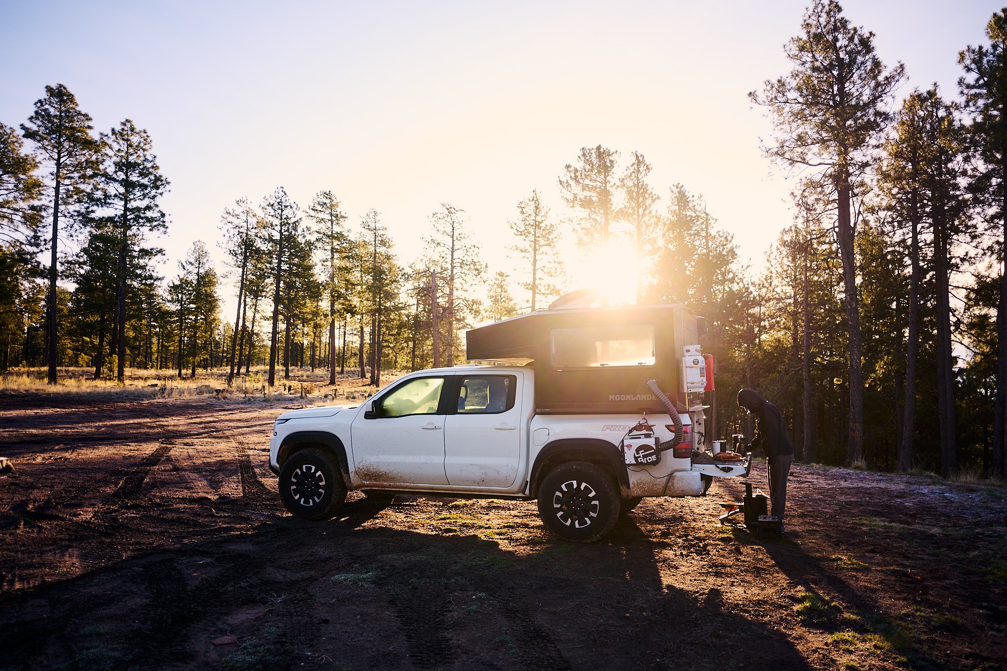 A white pickup truck parked on muddy ground in a forested area during sunset, with a large green camper attached to the truck and a person standing beside it.