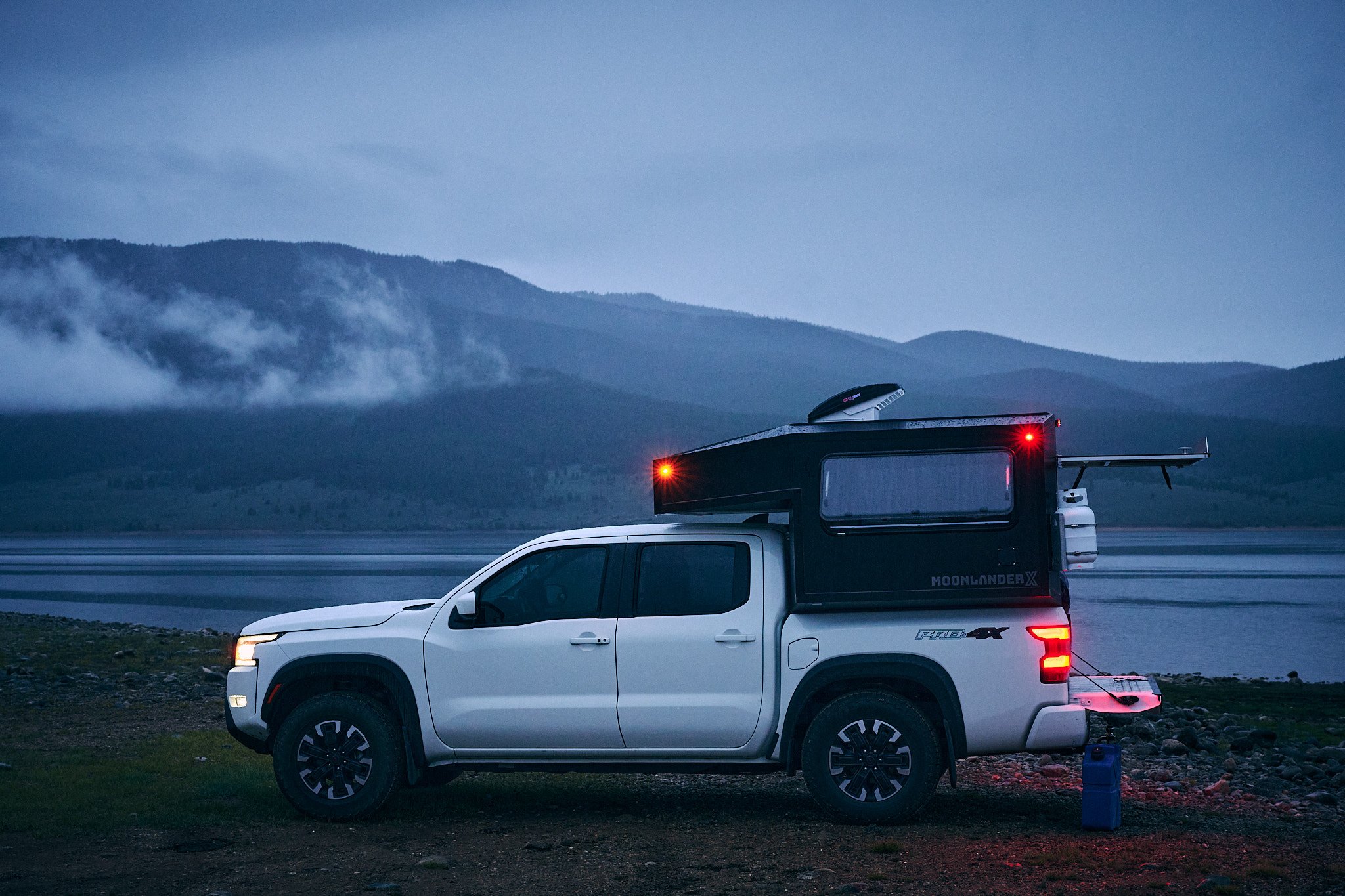 A white pickup truck with a camper shell parked on rocky terrain by a body of water, with mountains and clouds in the background during dusk or dawn.