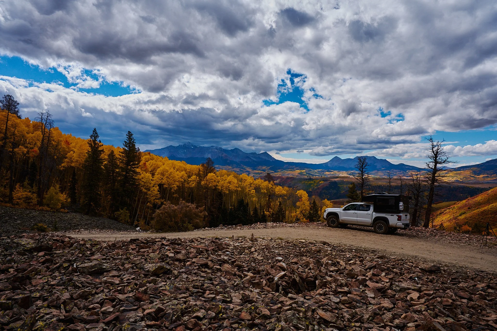 A scenic landscape with a partly cloudy sky, distant mountains, and trees displaying fall colors. A white pickup truck with a rooftop tent is parked on a dirt area in the foreground.