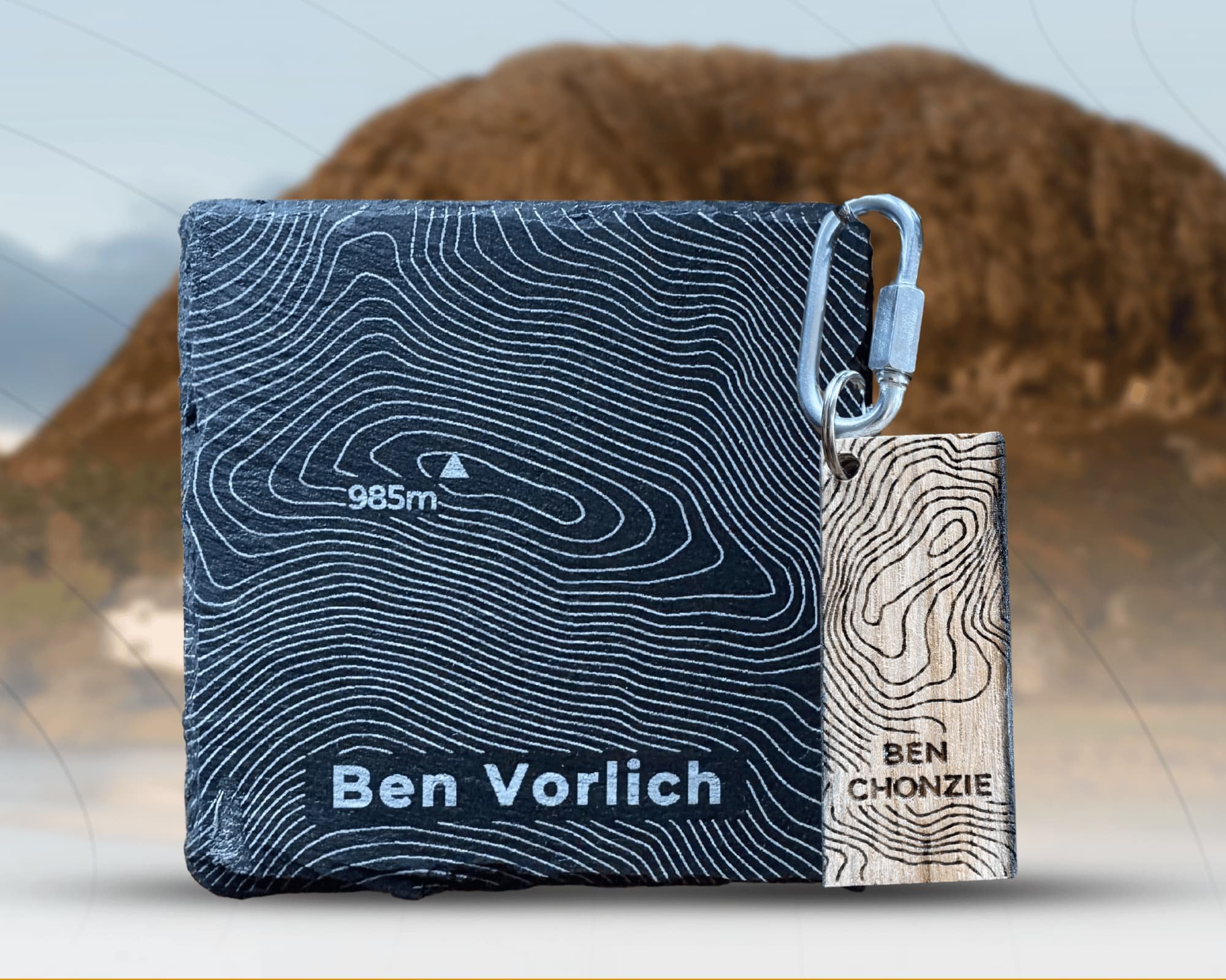A black and white topographic design pouch with the name Ben Vorlich and a mountain symbol, connected to a wooden tag with similar keyboard-like topography lines and the name Ben Chonzie, attached to a metal carabiner. A mountain in the blurred background.