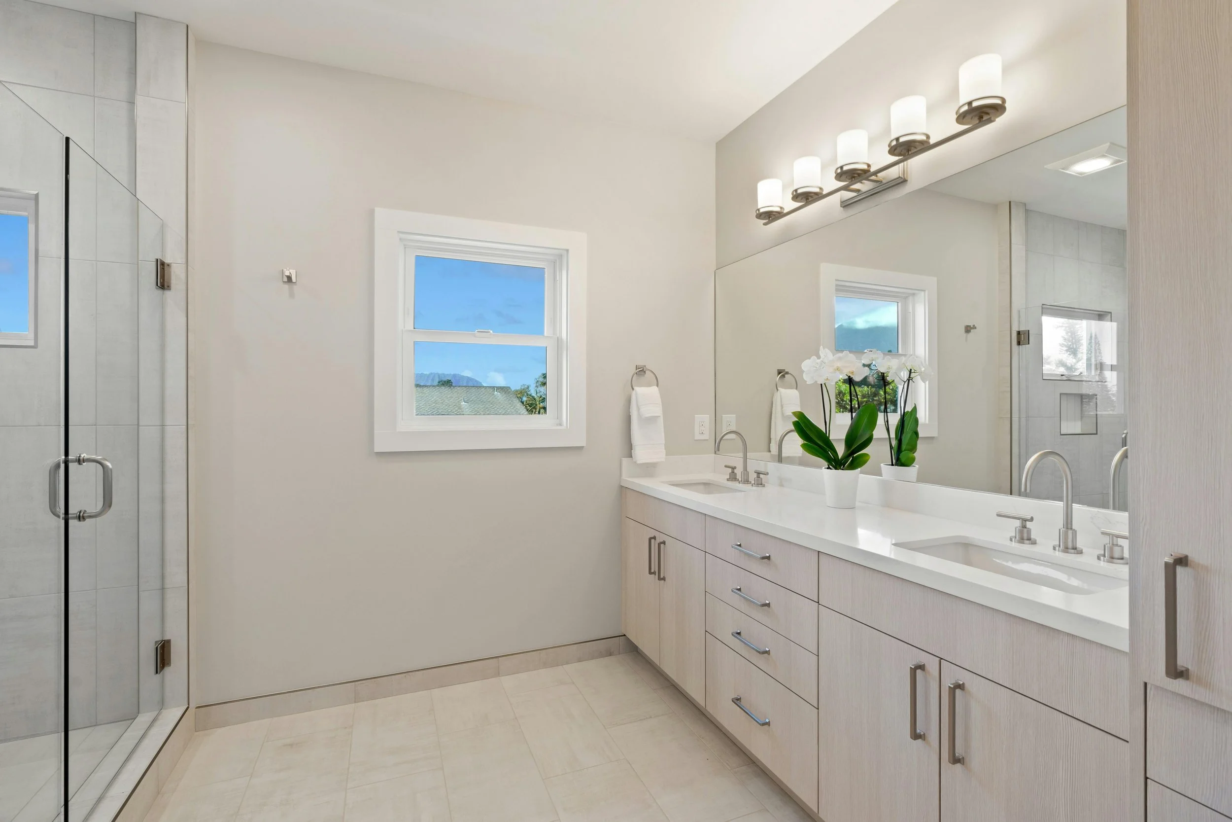 A modern bathroom with a double sink vanity, large mirror, white countertops, potted orchids, window with a view, and a glass shower enclosure.