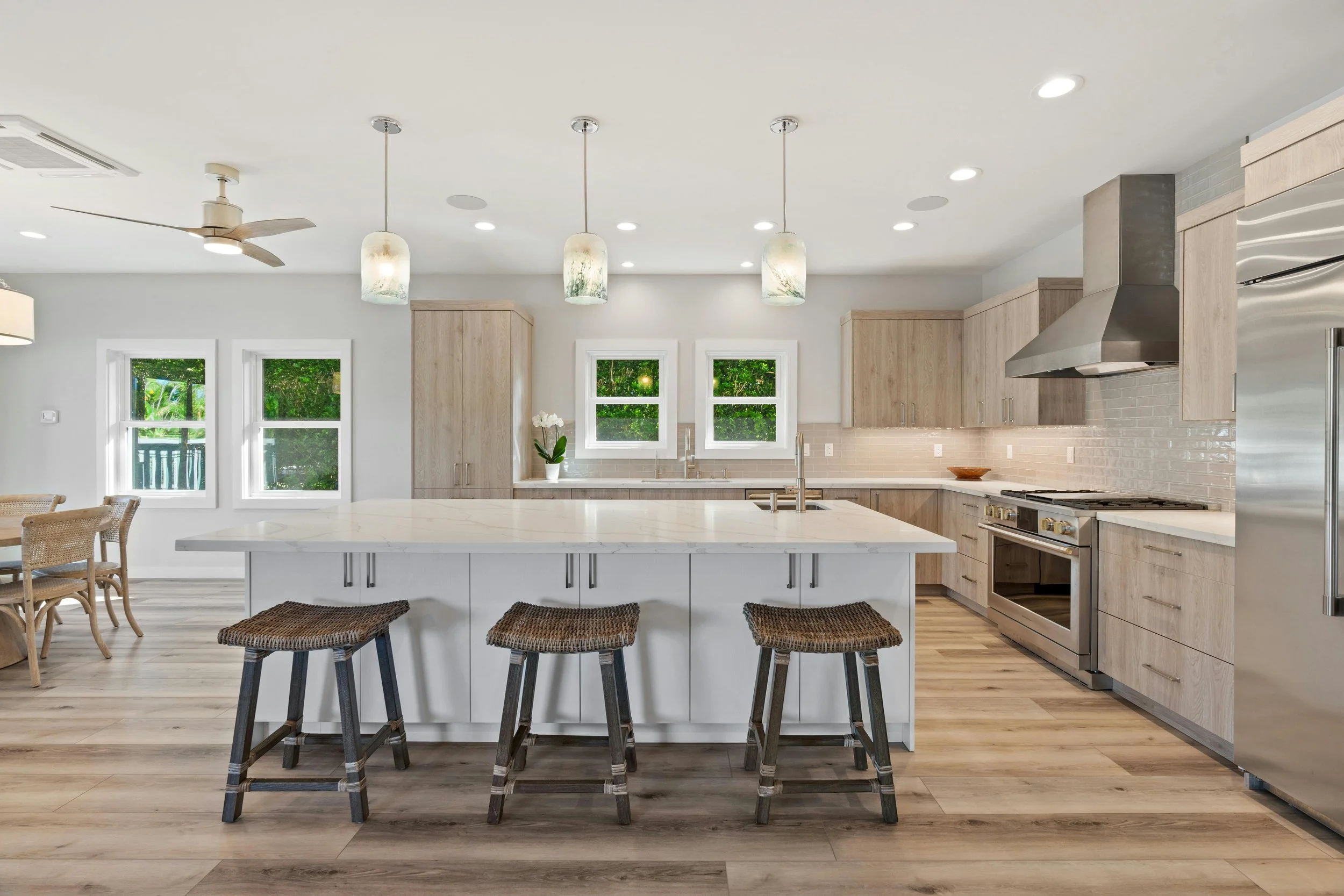 Modern kitchen with white and light wood cabinets, stainless steel appliances, a white island with bar stools, three pendant lights, and two windows showing greenery outside.