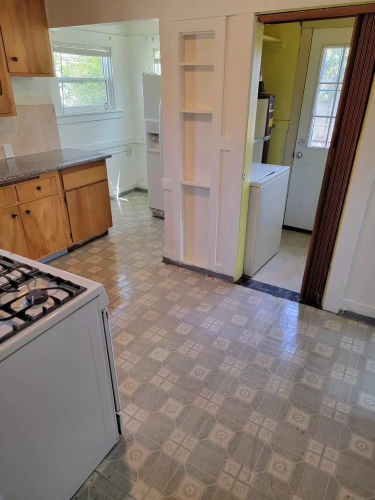 A kitchen with wooden cabinets, a white stove, and a window with natural light. Adjacent is a laundry area with a sink, washing machine, and a door leading outside. There's a small built-in shelf on a white sliding door.