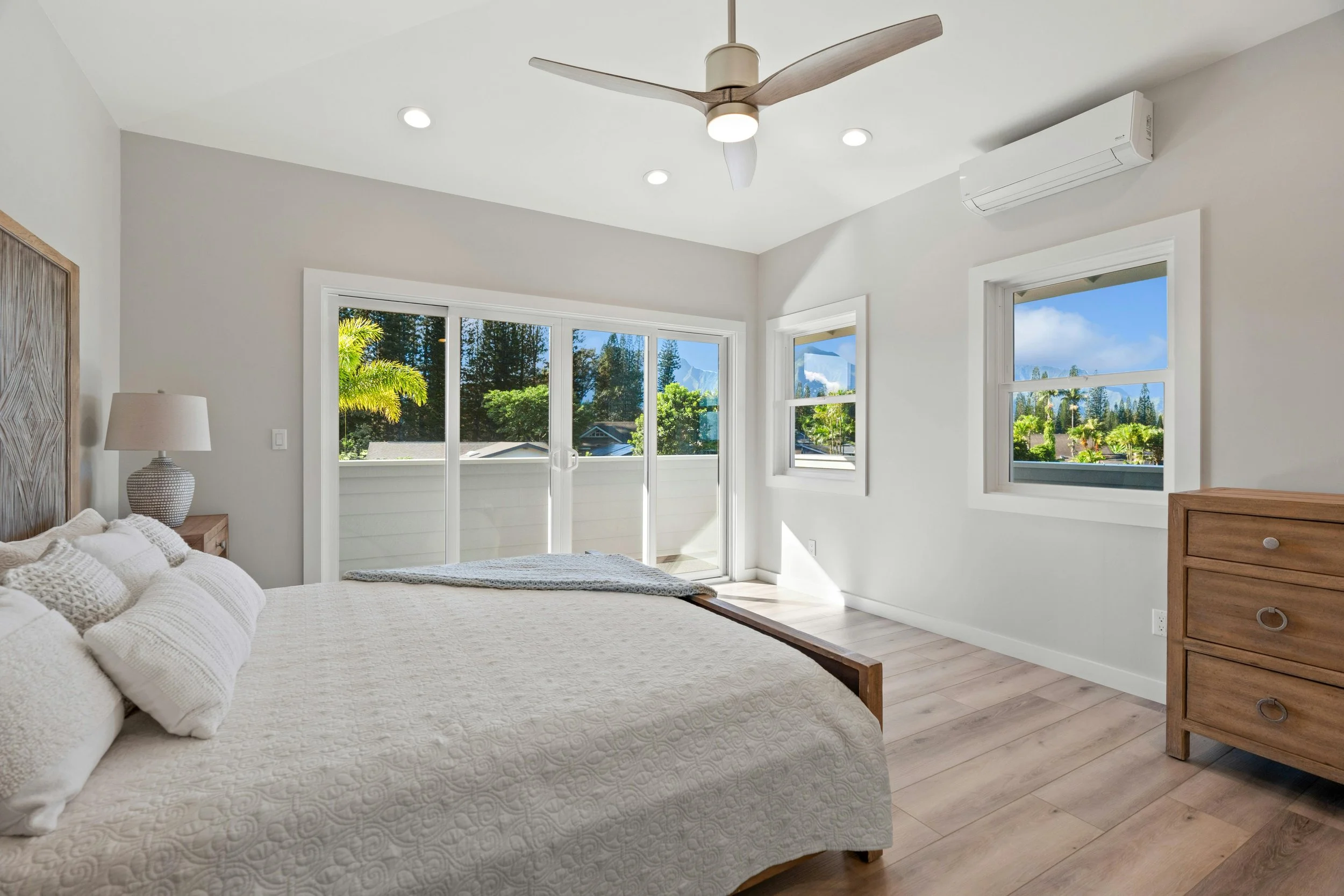 A bright bedroom with a large bed covered in white bedding and pillows, a wooden nightstand with a lamp, large sliding glass door and windows showing lush greenery outside, a ceiling fan with wooden blades, and a white air conditioning unit on the wa