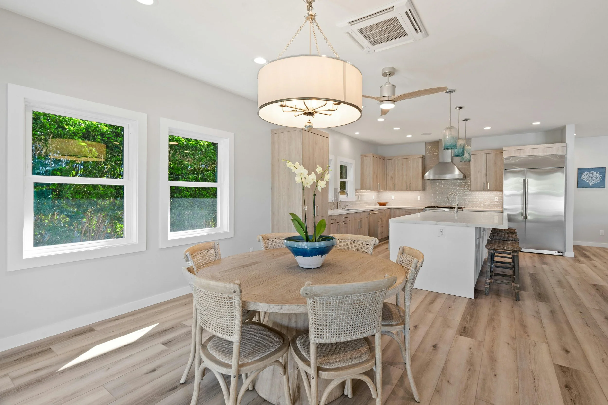 Bright, modern kitchen and dining area featuring a round wooden table with six wicker chairs, a white orchid plant in a blue and white pot, three large windows with greenery outside, light wooden cabinetry, a white kitchen island, and pendant lights 