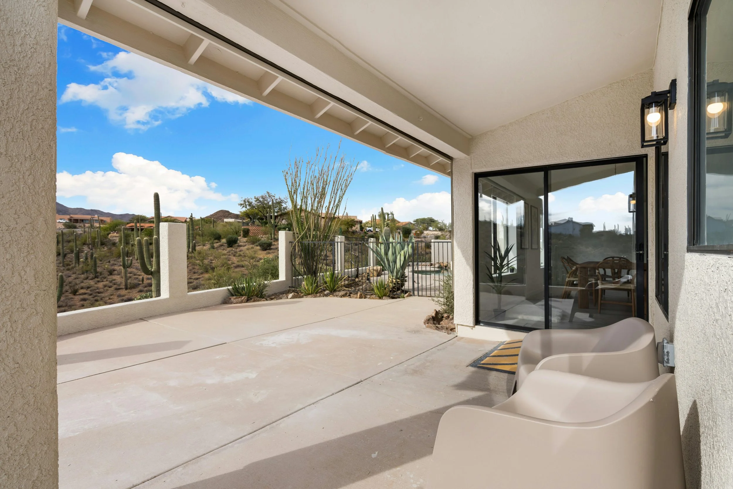 A covered outdoor patio area with two modern white chairs, large glass sliding doors, and desert landscape with cacti and hills in the background under a partly cloudy sky.