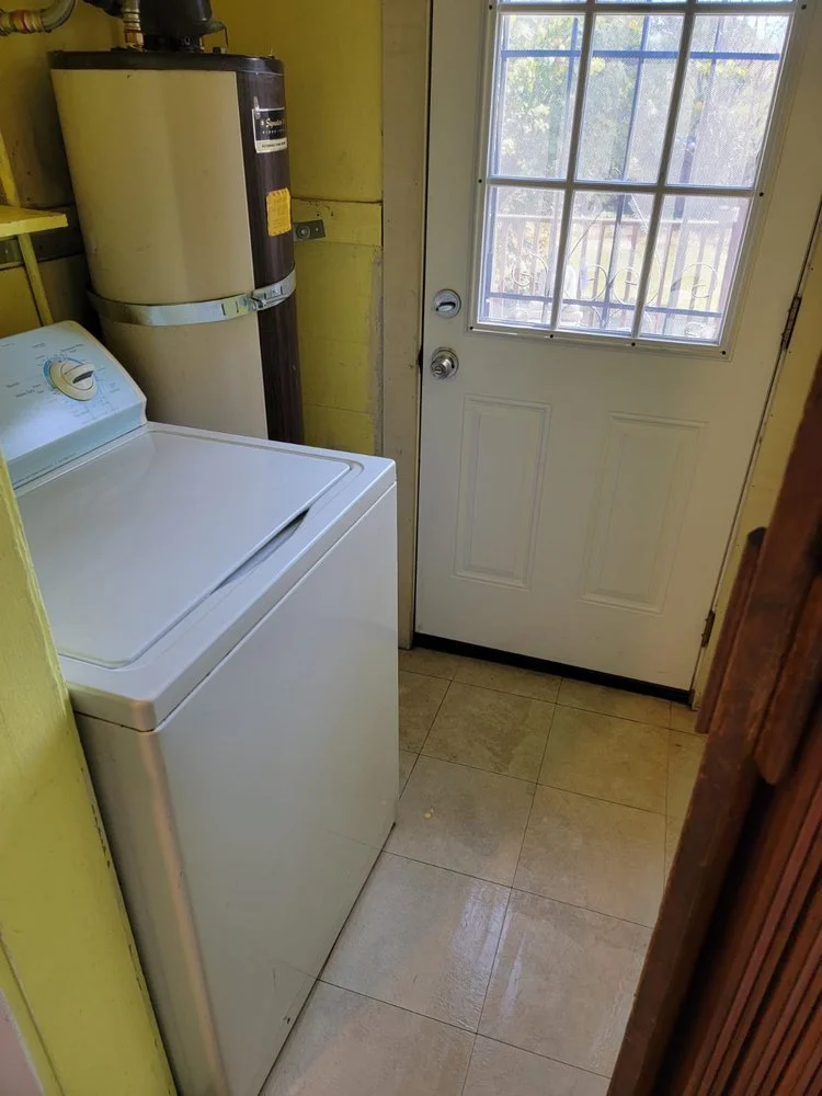 A small laundry room with a white top-loading washing machine, a yellow wall, and a door with a window leading outside. A water heater is mounted on the wall next to the machine.