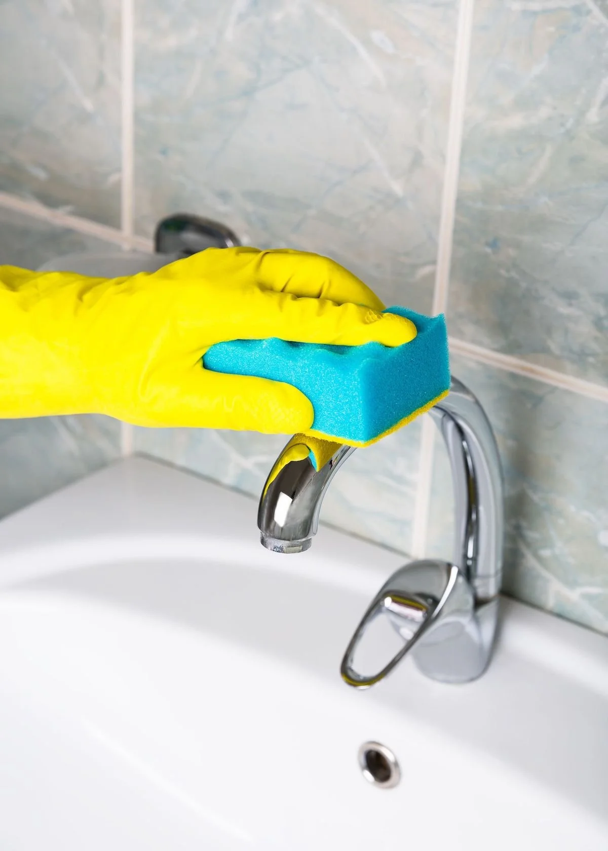 A person wearing yellow rubber gloves cleaning a bathroom sink with a blue sponge.