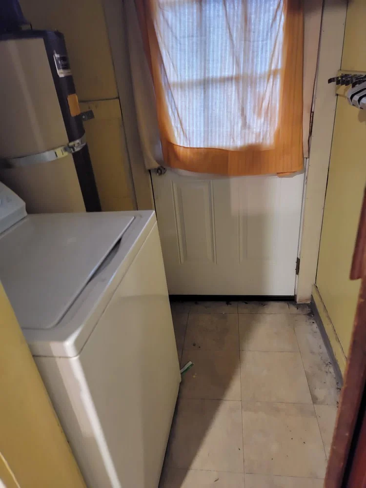 A small laundry room with a white washing machine, beige walls, a door with a window covered by an orange curtain, and a tiled floor with some dirt or wear near the door.