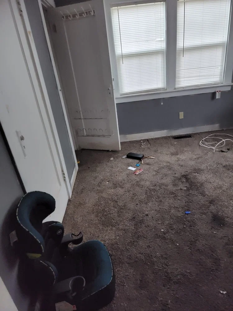 Empty room with a car seat on a brown carpet, large windows with blinds, open closet door, some scattered items on the floor including a remote and small cards, and electrical wires.