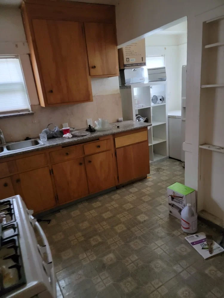 Empty kitchen with wooden cabinets, a sink, some cleaning supplies, an open shelving unit, and a box on the floor.