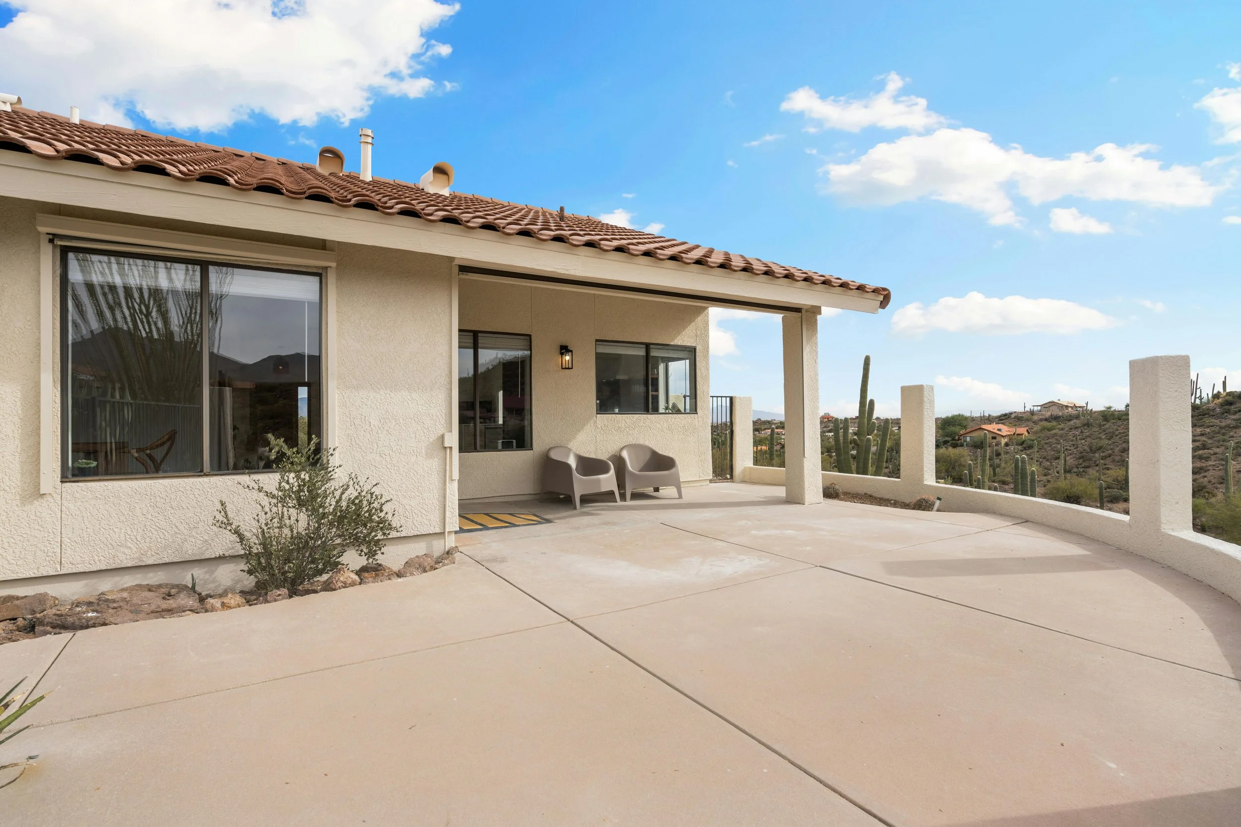 View of a house with a terracotta tiled roof, large glass sliding doors, an outdoor patio area with two chairs, and desert landscape with cacti in the background.