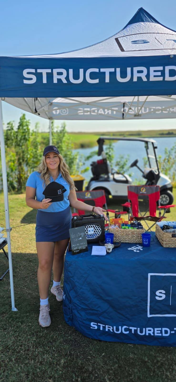 A young woman standing next to a table at an outdoor event, smiling and holding a black cap. The table has various items and a sign, with a golf cart and a scenic lake view in the background.