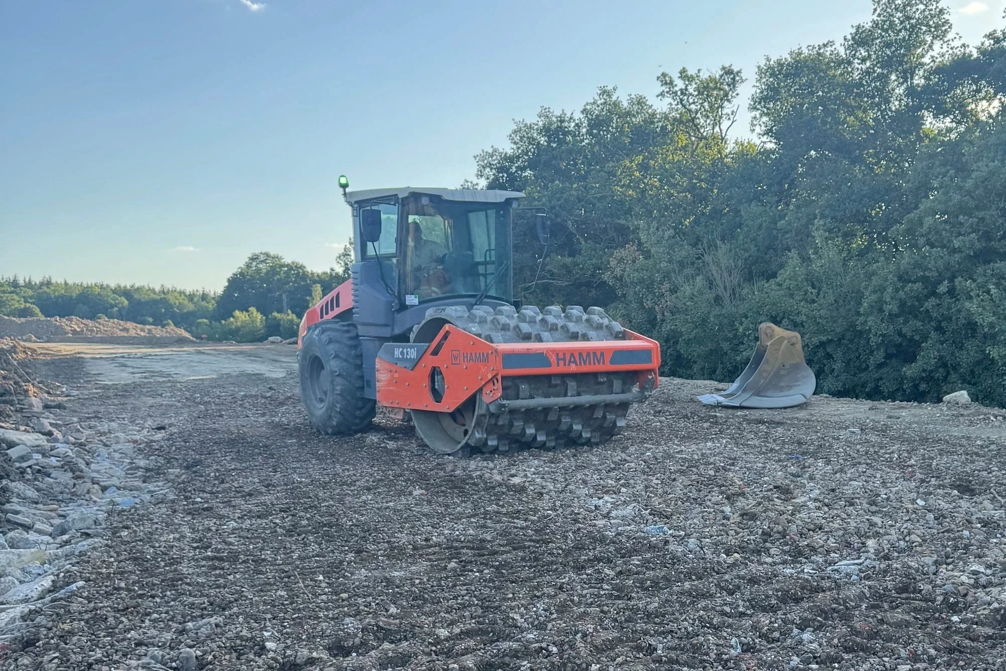 Construction site with a small orange and black HAMM road roller on dirt ground, surrounded by sparse trees and bushes.
