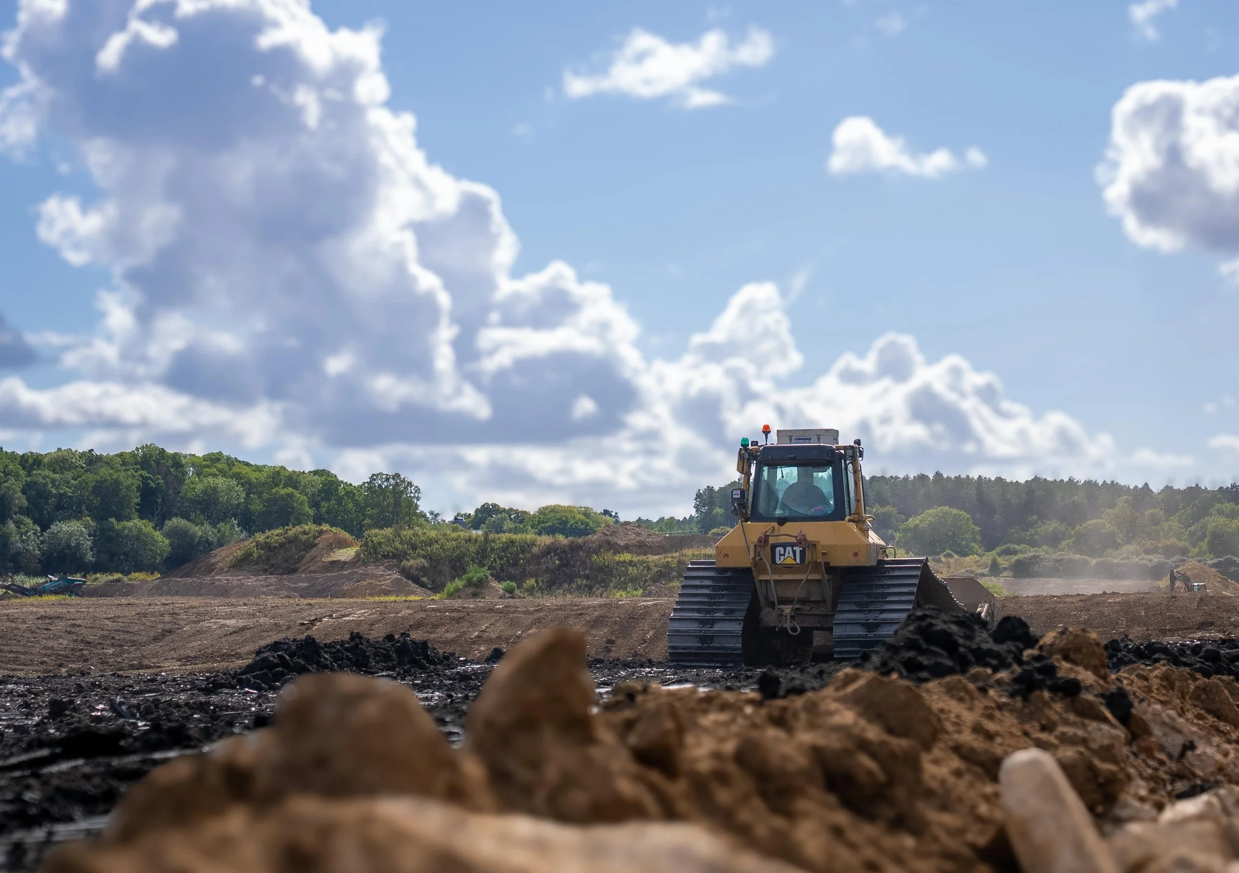 A yellow bulldozer on a Landfill site with green trees in the background and a partly cloudy sky.