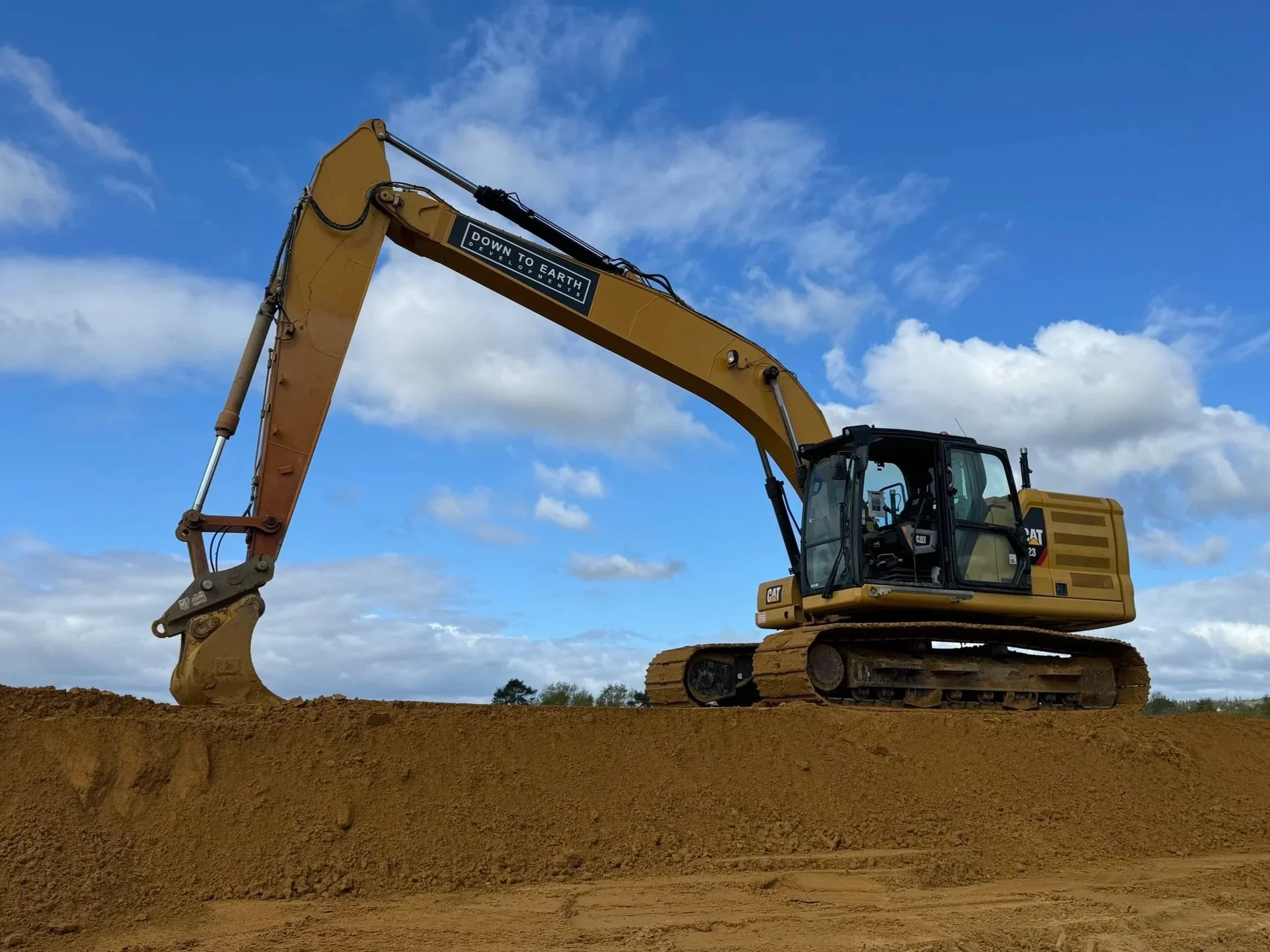 A yellow excavator with a black and white sticker that reads 'Down to Earth' is on a mound of dirt against a partly cloudy sky.