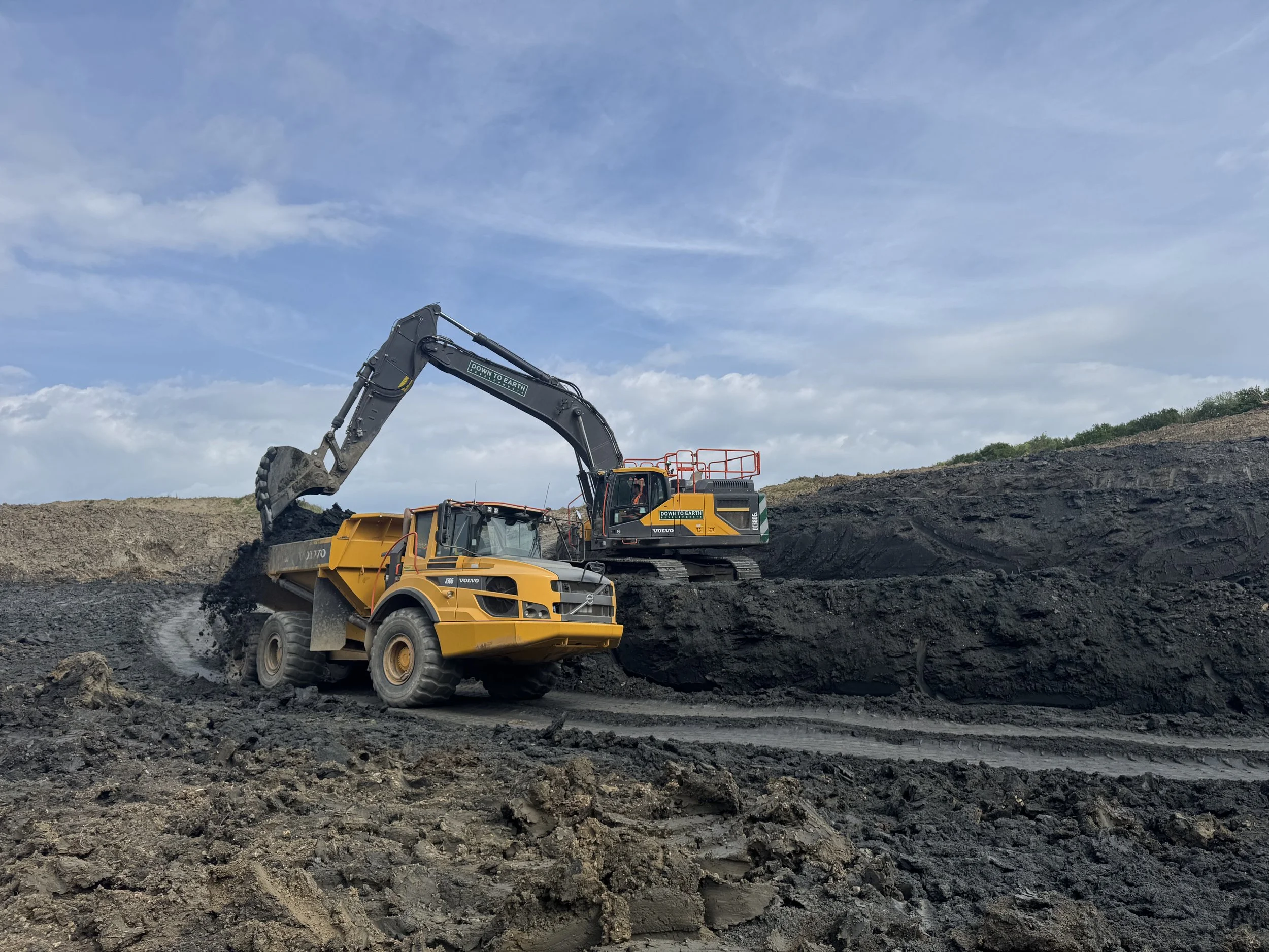 An excavator and a dump truck working together on a construction site or mining area under a partly cloudy sky.