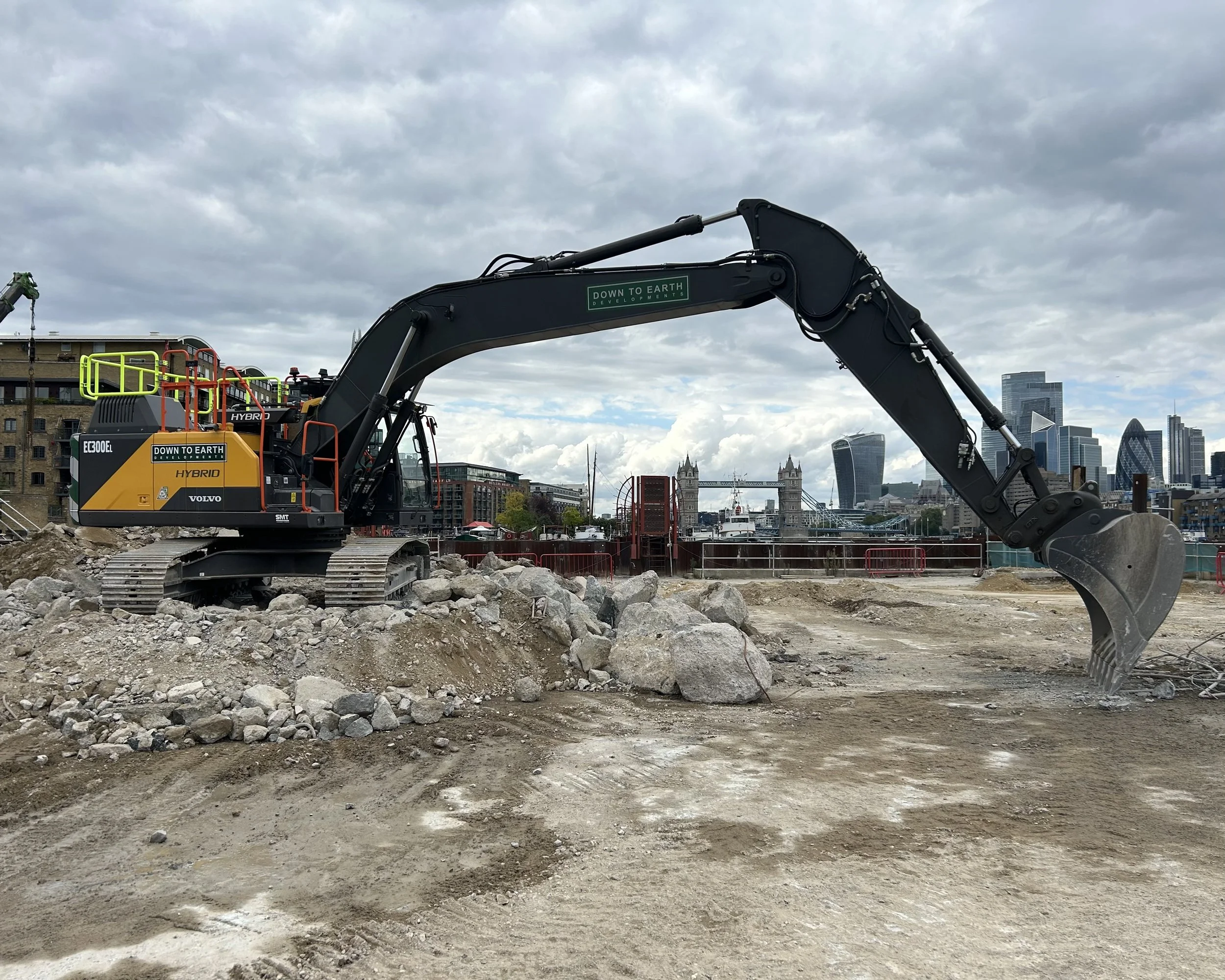 Construction excavator digging up dirt and rocks near a city riverfront with modern skyscrapers and a cloudy sky in the background.